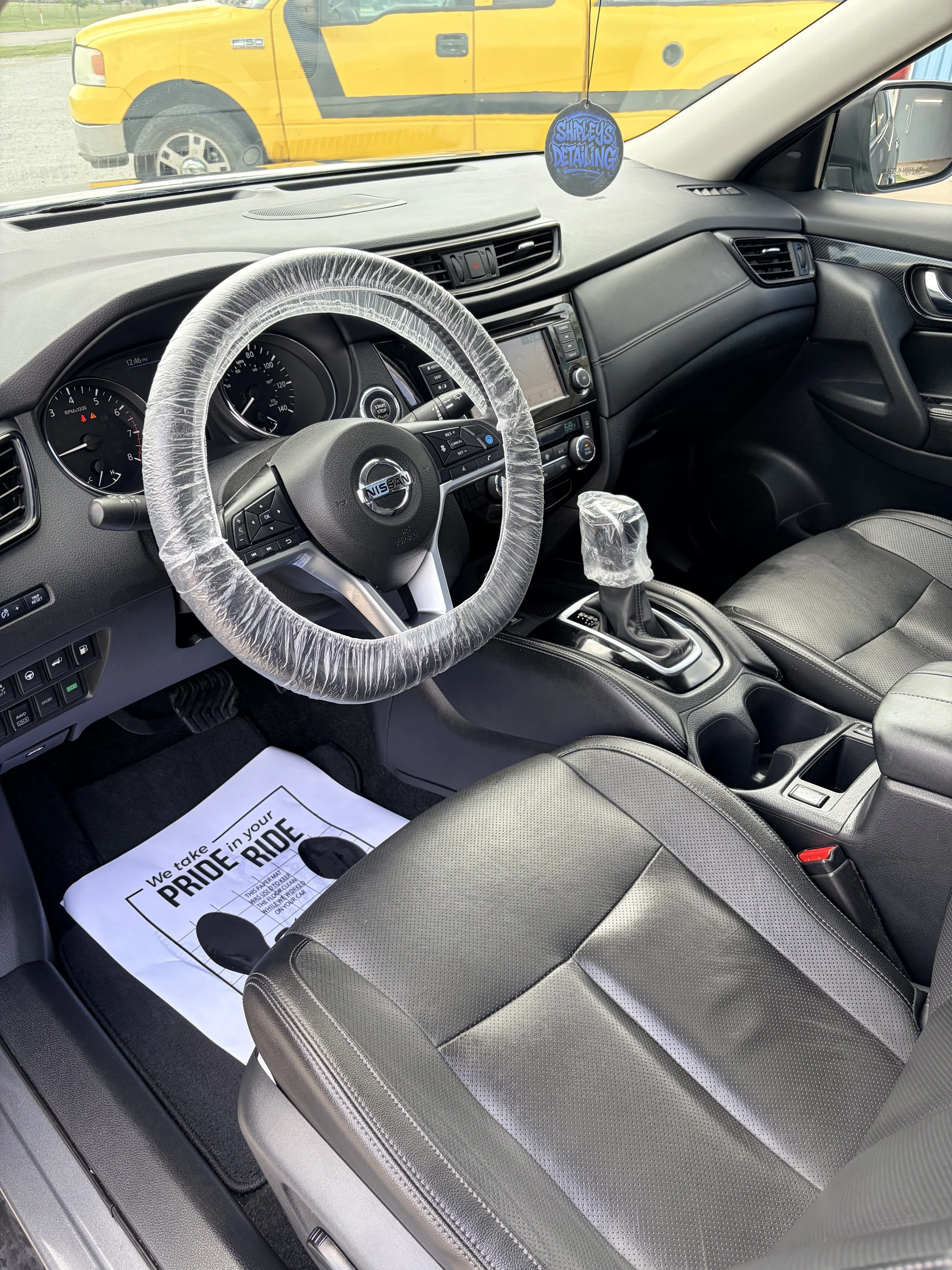 The interior of a Nissan vehicle with a plastic wrapped steering wheel and gear shift, a dashboard with a navigation screen, and a front seat with a paper mat that reads 'We take pride in your ride'.