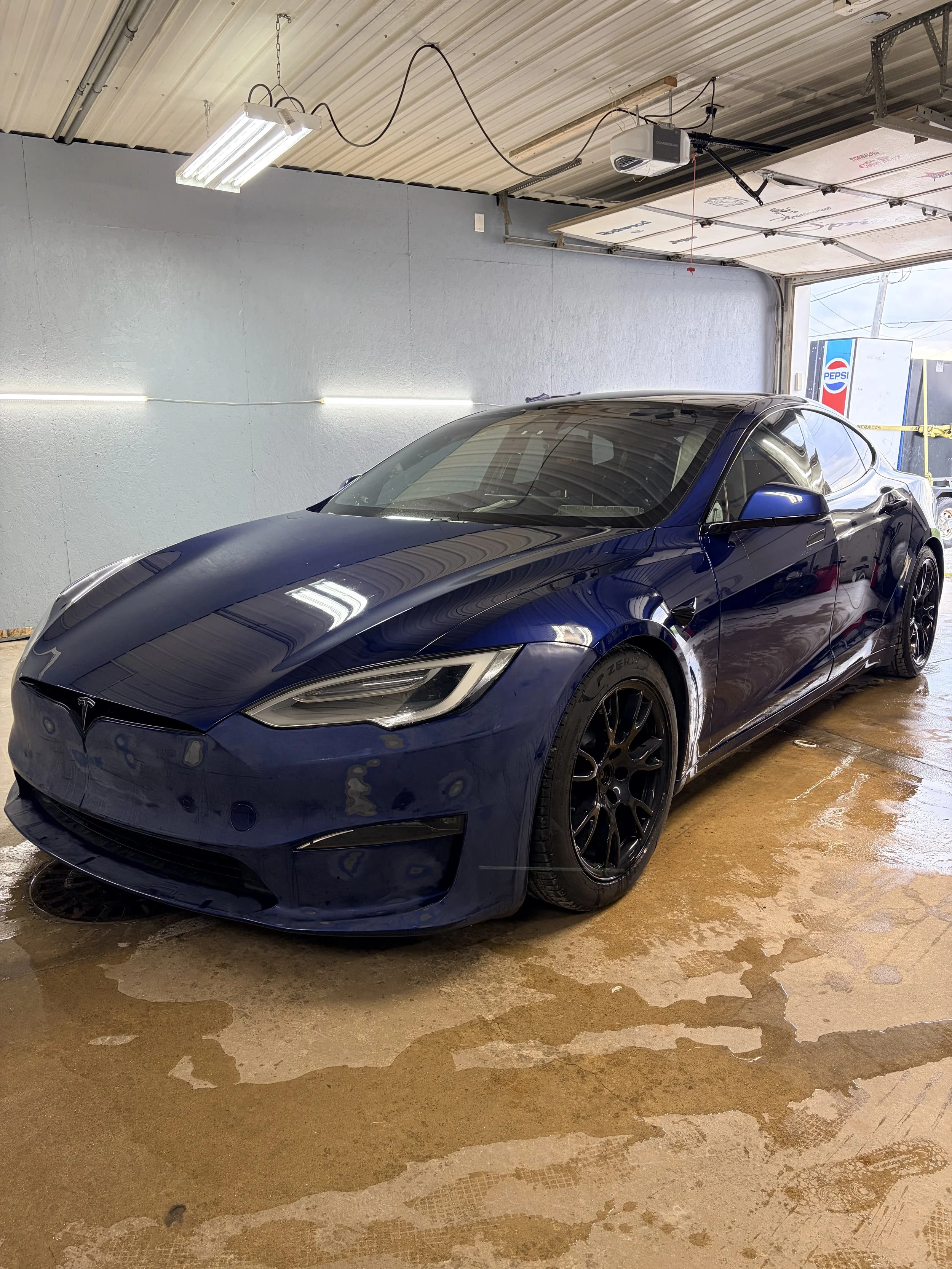 A dark blue Tesla sports car parked inside a garage with a concrete floor and a white wall. The garage door is open, revealing a Pepsi vending machine outside. The garage ceiling has fluorescent lighting and visible wiring.