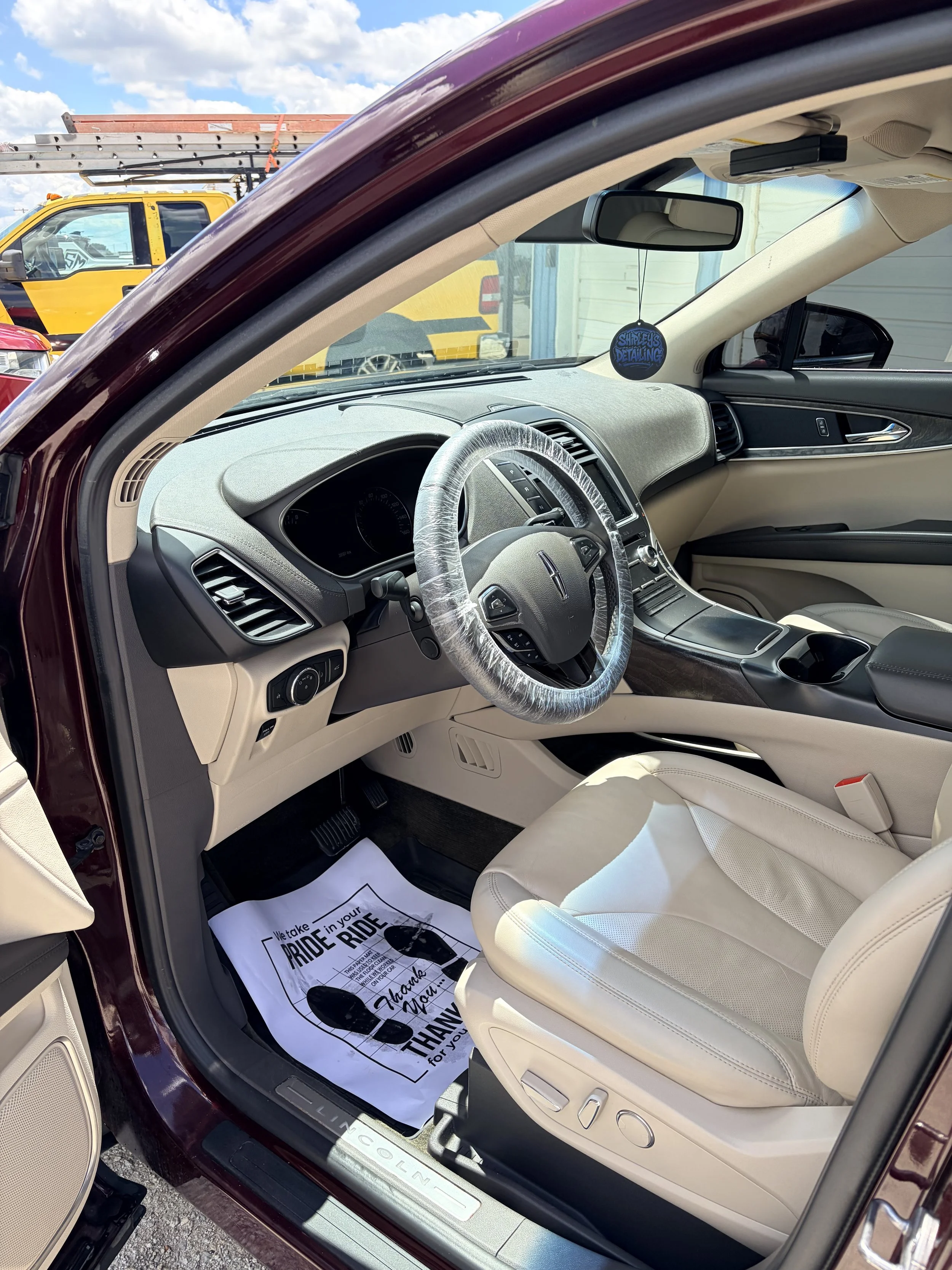 Interior of a Lincoln vehicle, showing the beige leather seat, steering wheel with a plastic cover, and dashboard. Outside the vehicle, other cars and a yellow truck with a ladder truck are visible. A black circular air freshener hanging from the rea