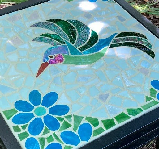Glass mosaic table featuring an iridescent hummingbird with flowers. 18 x 18 x 20 inch. Commissioned in 2026.