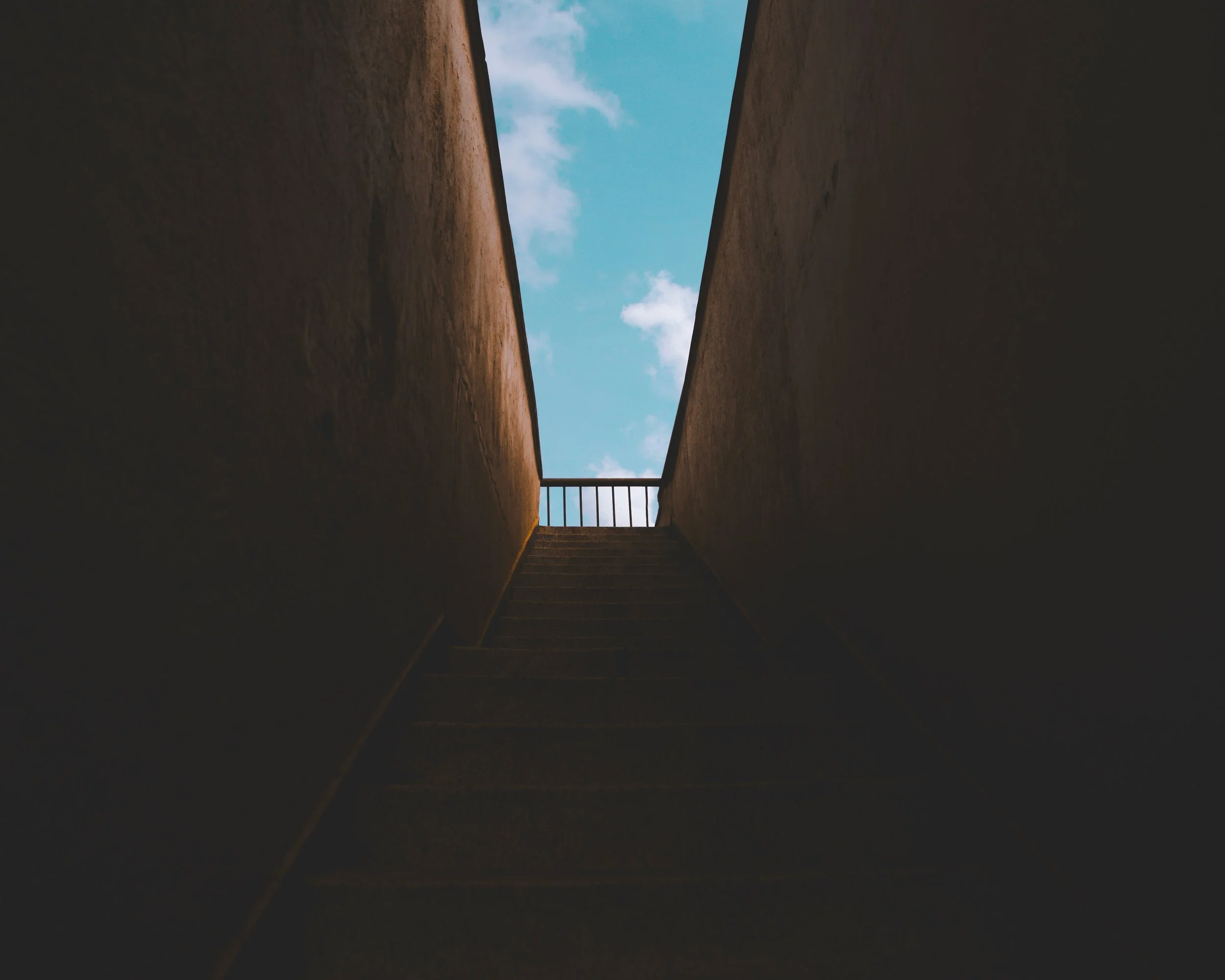 moody colour photo of the view looking up steps form a tunnel or bridge up to the blue sky