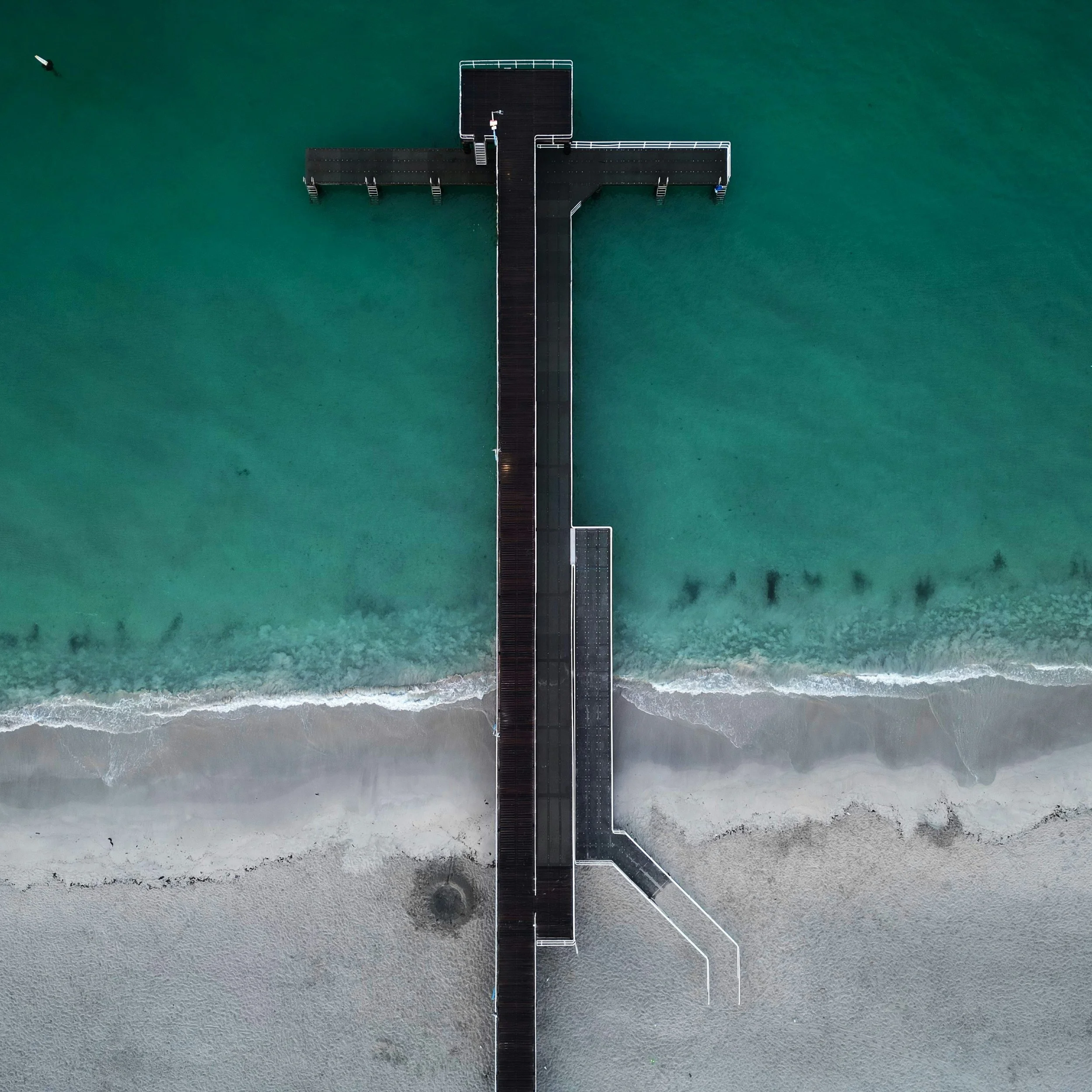 colour photo taken form above of a jetty or pier jutting out of the sand into turquoise sea
