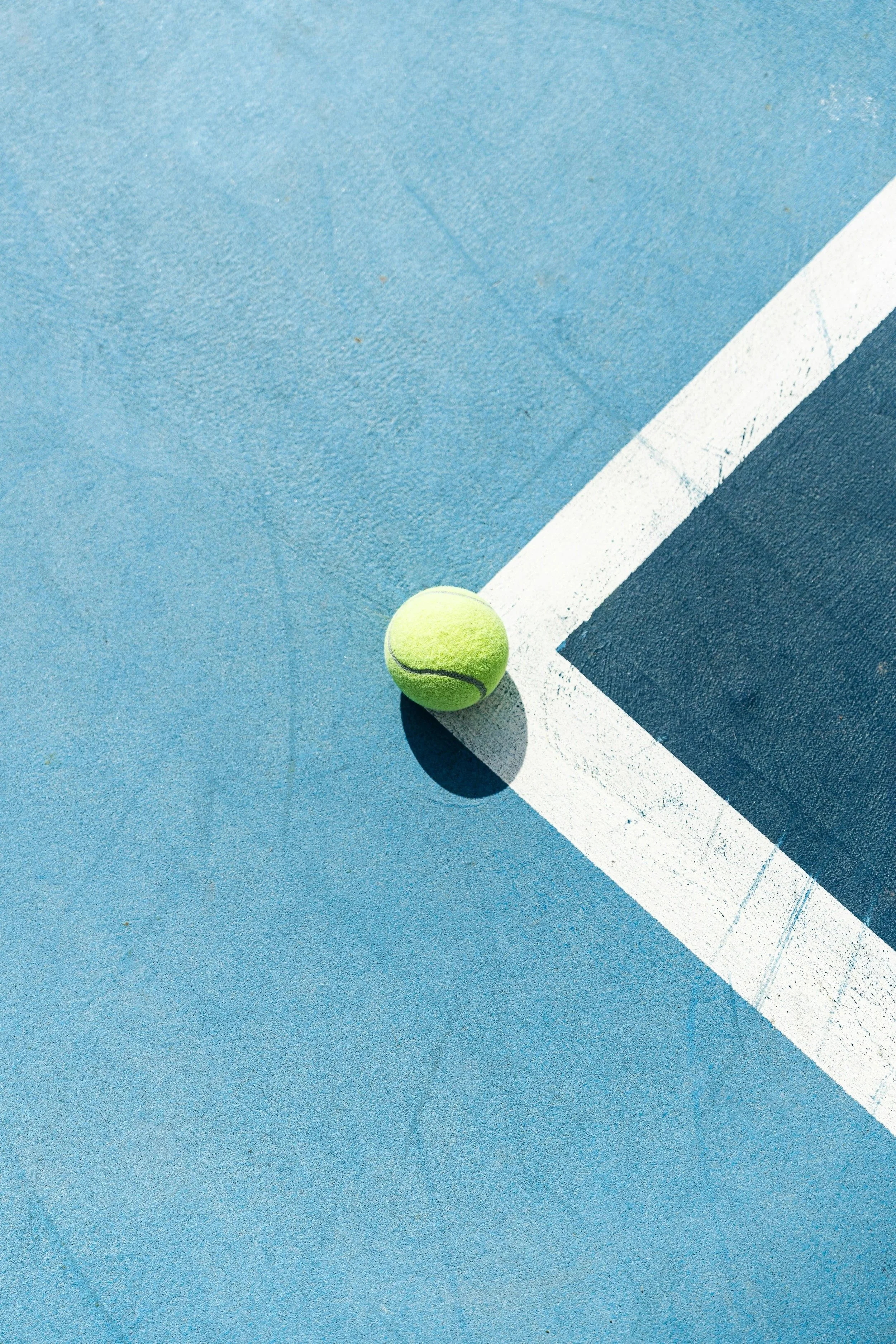 colour photo of a tennis ball on the point of a tennis court line, in blue tones