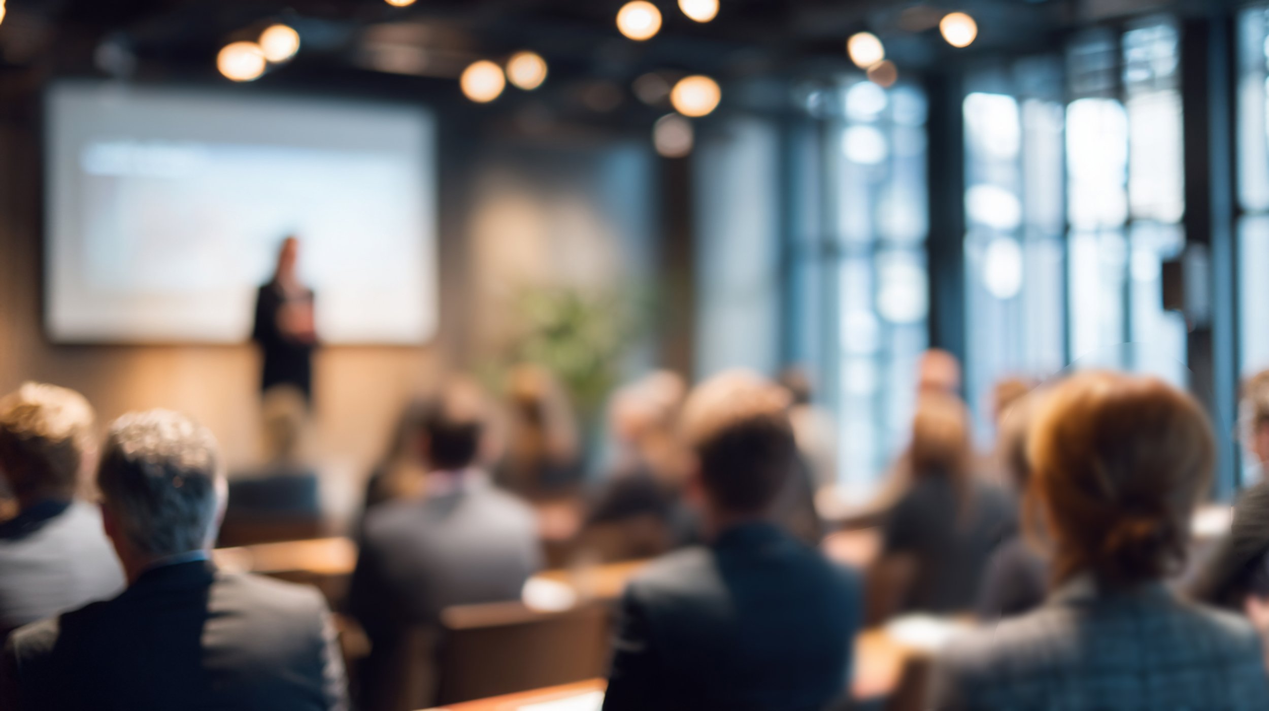 A blurred image showing an audience attending a presentation or lecture in a modern, well-lit conference room with large windows, a large screen at the front, and a speaker at the podium.