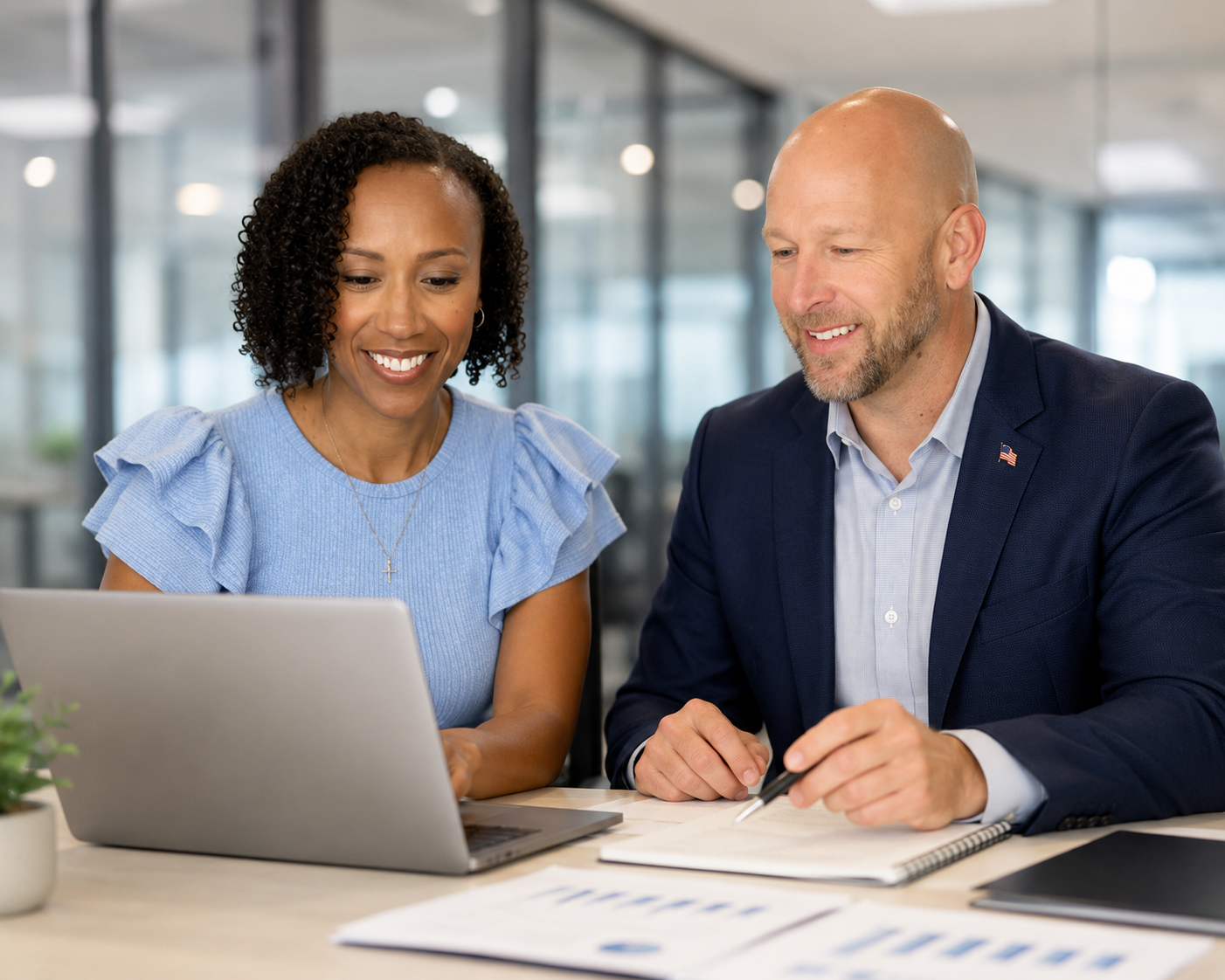Two professionals, a woman and a man, are sitting at a desk looking at a laptop and smiling in an office setting.