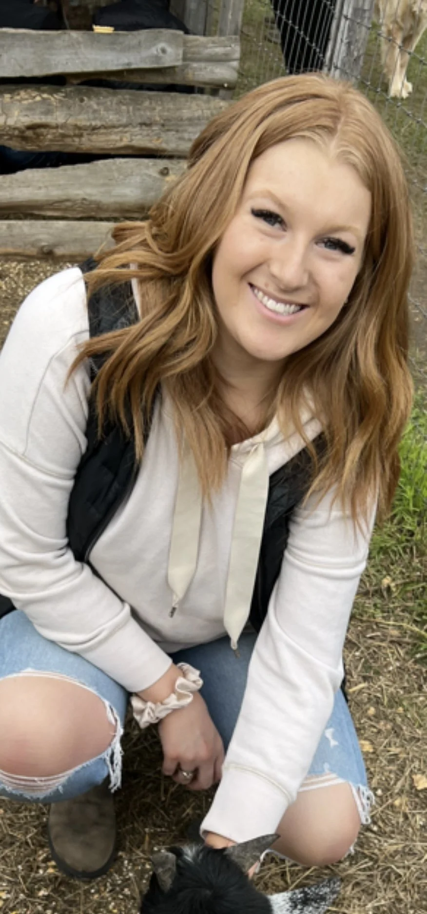 A smiling woman with long red hair, wearing a white hoodie, ripped jeans, and gray boots, is crouching outdoors near a black and white goat with a background of wooden logs and a wire fence with animals.