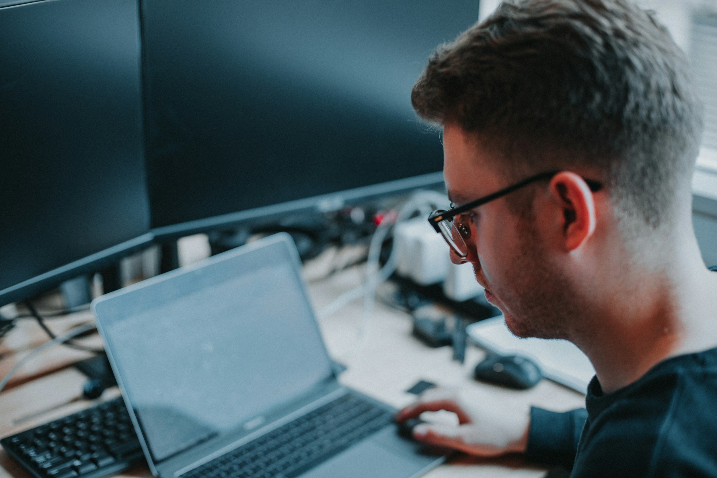 A person with short hair and glasses working on a laptop in a room with multiple monitors and electronic equipment.