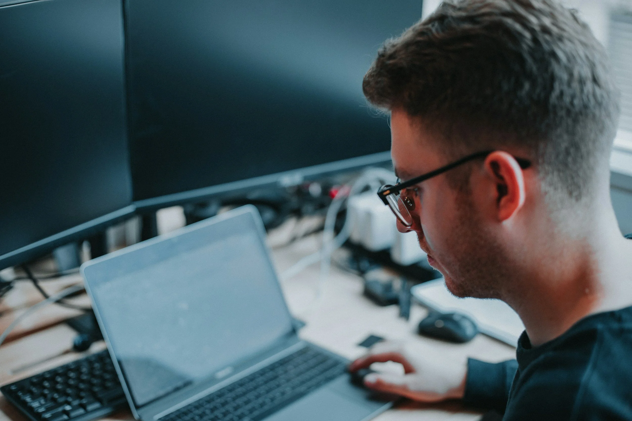 A person wearing glasses working on a laptop at a desk with multiple computer monitors.
