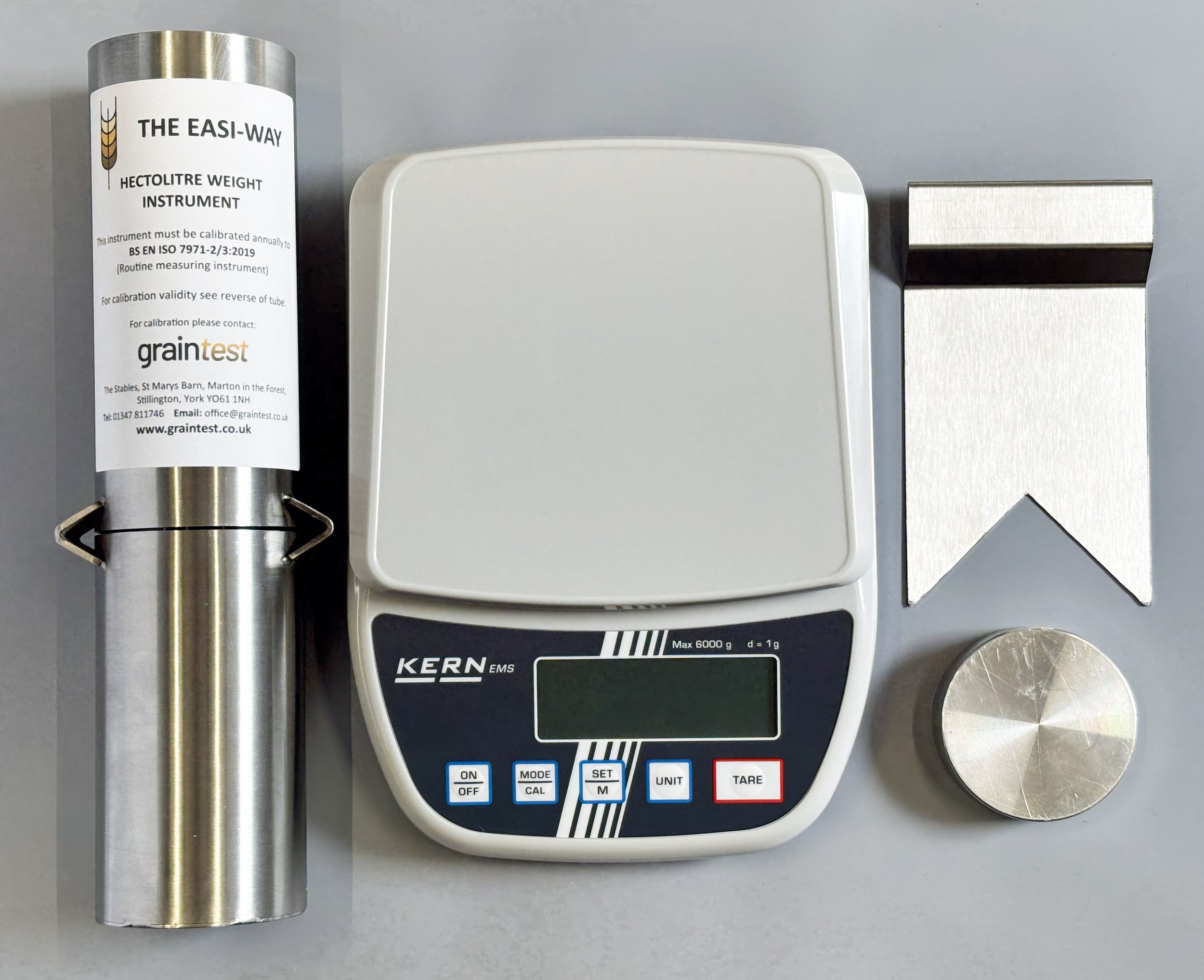 A metal tube, a white electronic digital scale, and a small metallic weight are arranged on a flat surface. The scale has several buttons with labels such as ON/OFF, MODE/CAL, SET/M, UNIT, and TARE.