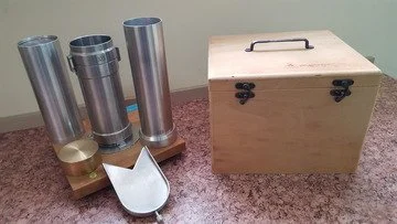 Set of three metal salt or pepper shakers with a wooden base, a wooden storage box with a handle, and a metal scoop on a pink speckled countertop.