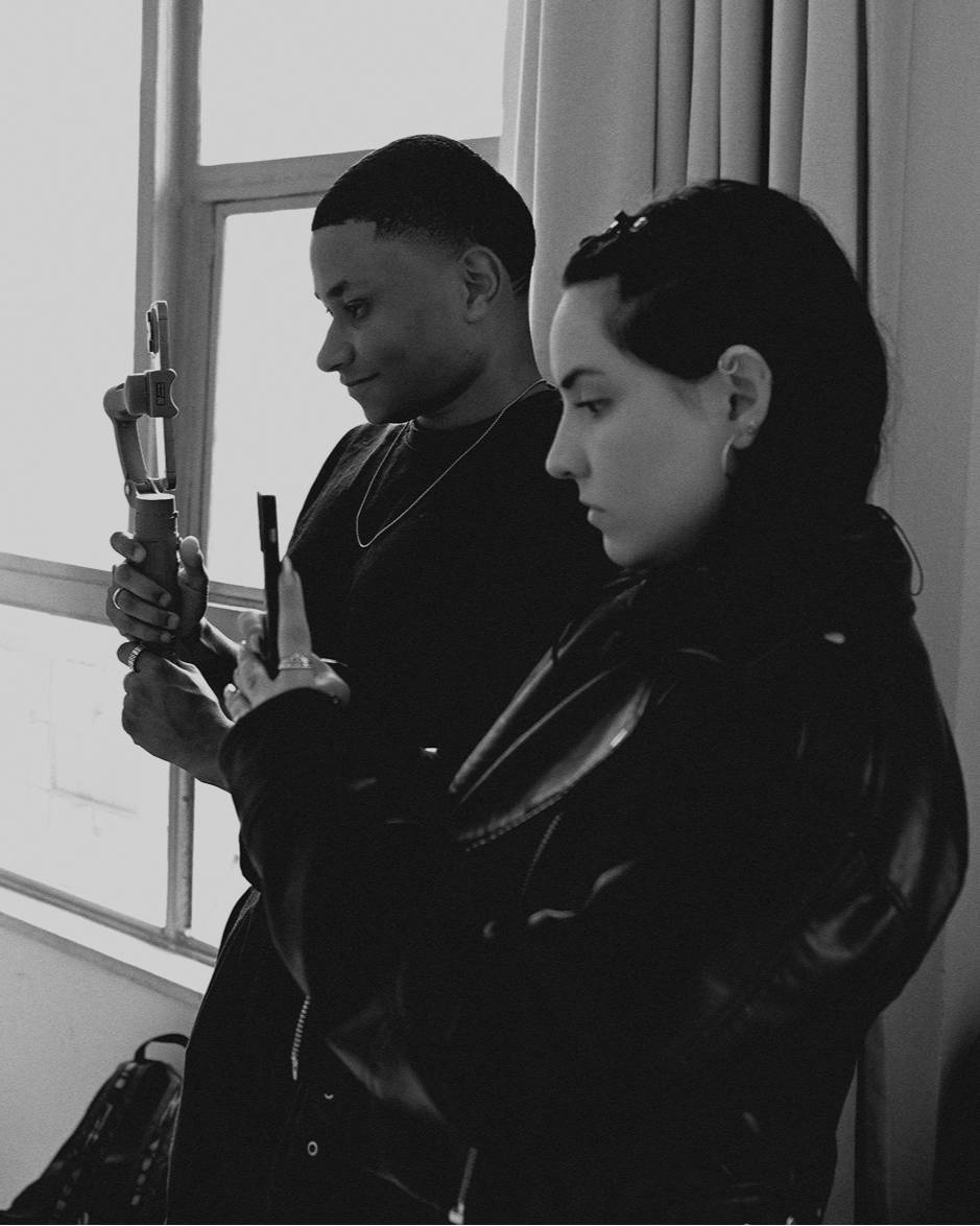 Dois jovens usando roupas pretas e acessórios, cada um filmando com celular, estão próximos a uma janela em uma sala com cortinas. A imagem é em preto e branco.