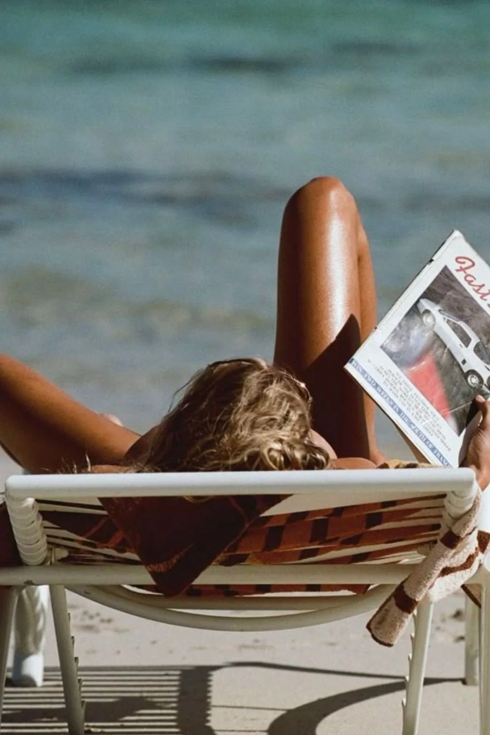 Woman relaxing on a lounge chair at the beach, reading a newspaper, with water in the background.