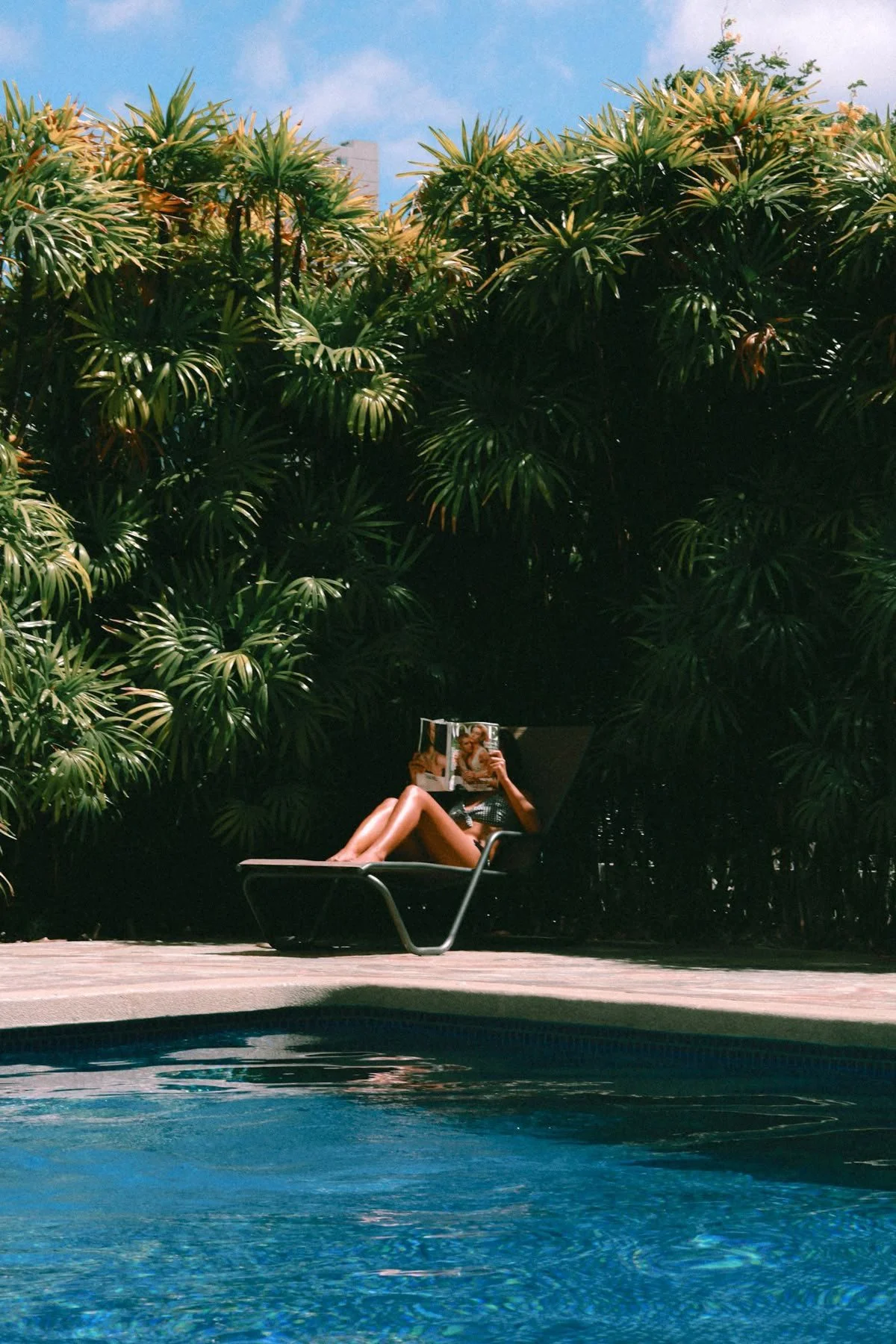 A woman relaxing on a lounge chair by a pool, reading a magazine, with lush green tropical plants and a blue sky in the background.