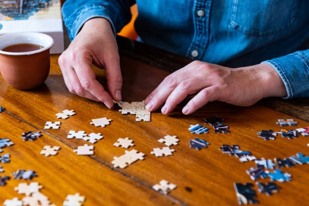 Person in a blue shirt working on assembling a jigsaw puzzle on a wooden table, with a coffee cup nearby.