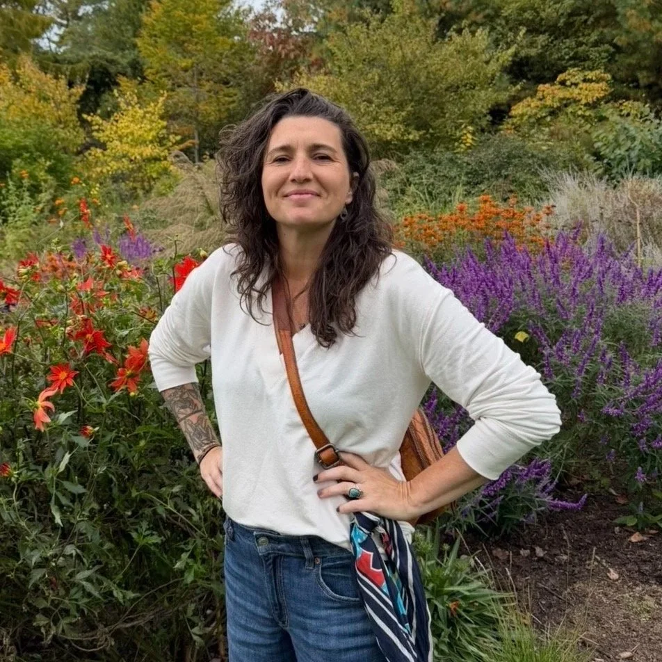 A woman with dark curly hair and tattoos on her left arm, wearing a white long-sleeve top, standing outdoors in a colorful garden with red, purple, and orange flowers and green trees in the background.