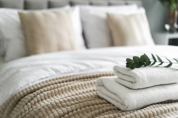 Neatly folded white towels with green leaves on a bed with white and beige bedding and pillows.