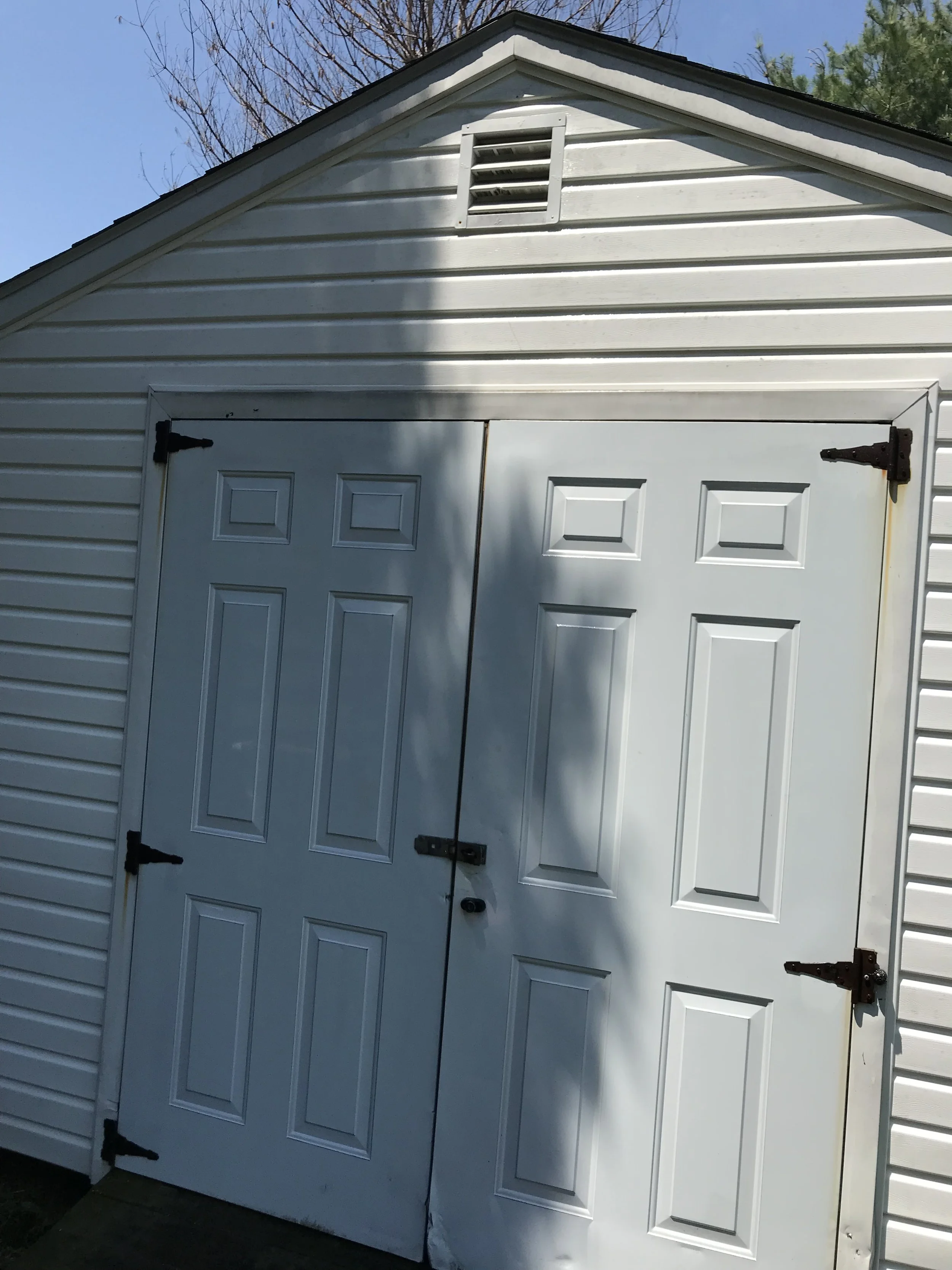 White garage door with black hinges and lock, part of a white shed with siding and a vent near the roof, under a blue sky with some tree branches in the background.