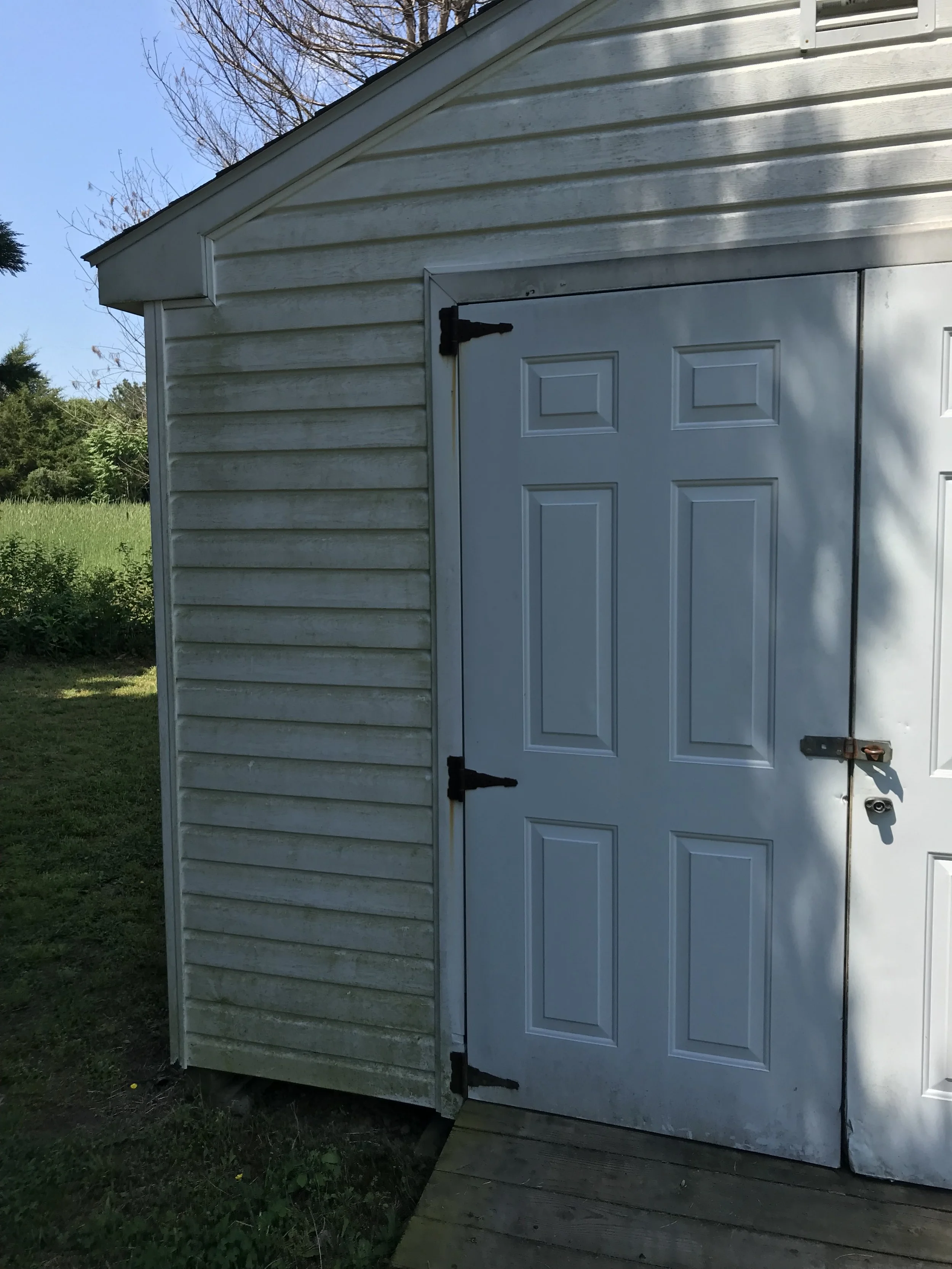 White shed with double doors and metal hinges, located on a grassy lawn with trees in the background. The shed has siding and a sloped roof.