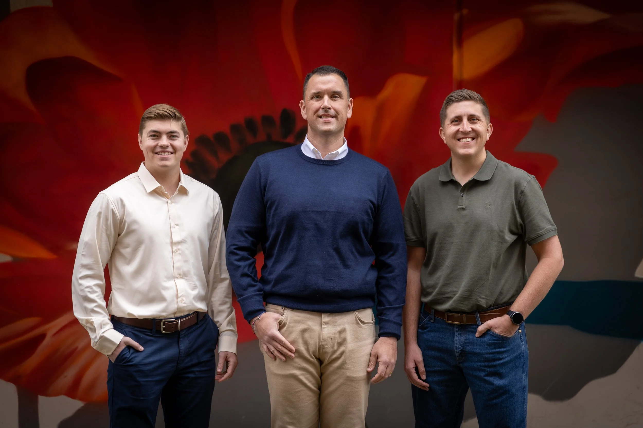Three men standing side by side, smiling in front of a vibrant, colorful background with red, orange, and yellow hues.