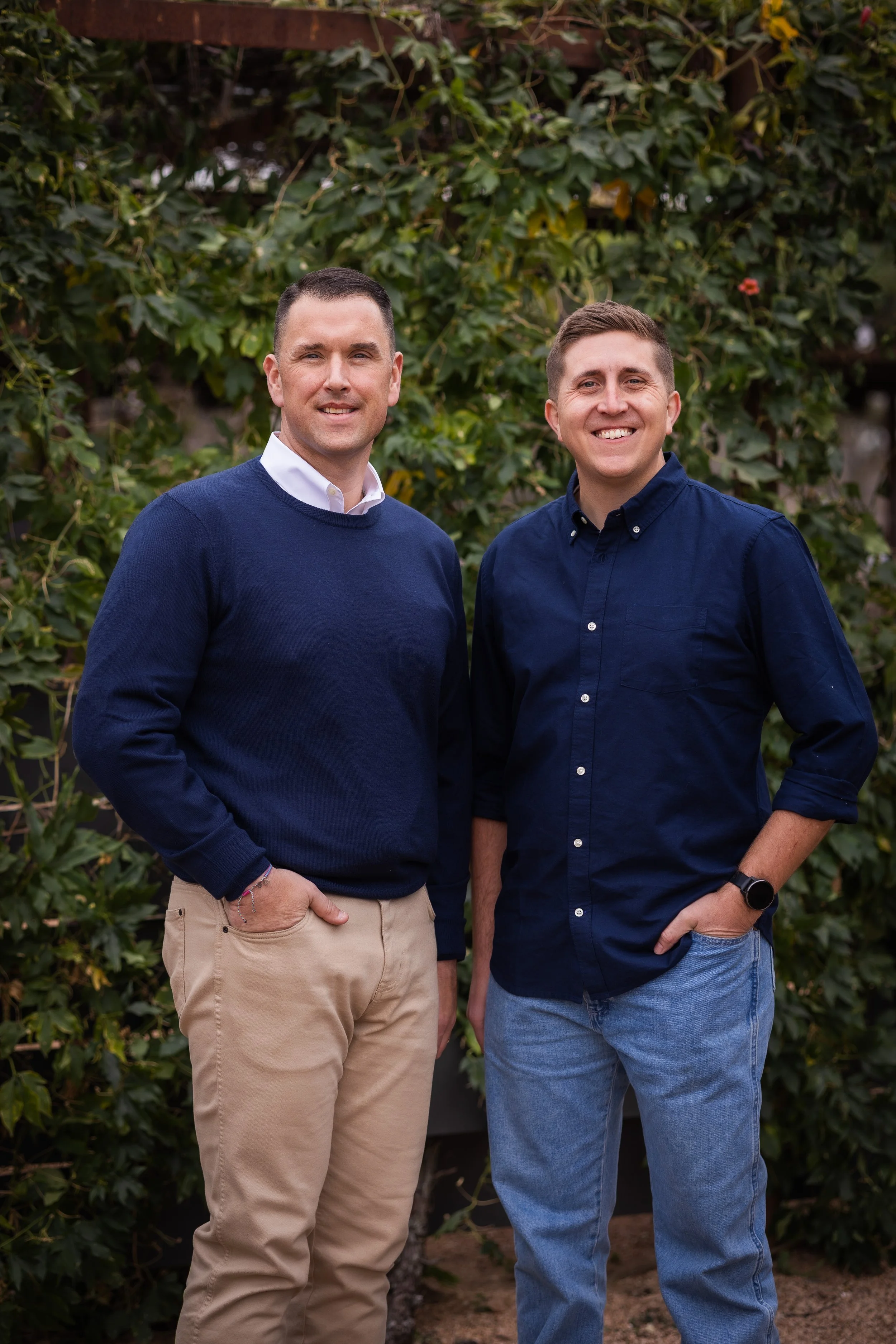 Two smiling men standing outdoors in front of greenery, wearing casual clothing with one in a navy sweater and khaki pants, the other in a navy shirt and blue jeans.