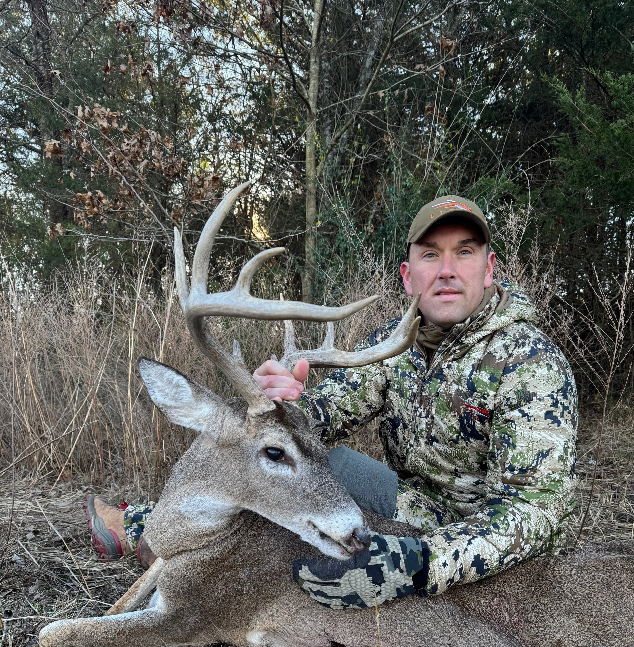 A man dressed in camouflage clothing sitting on the ground outdoors with a large deer that he has seemingly hunted. The man is wearing a cap and is surrounded by tall grass and trees.
