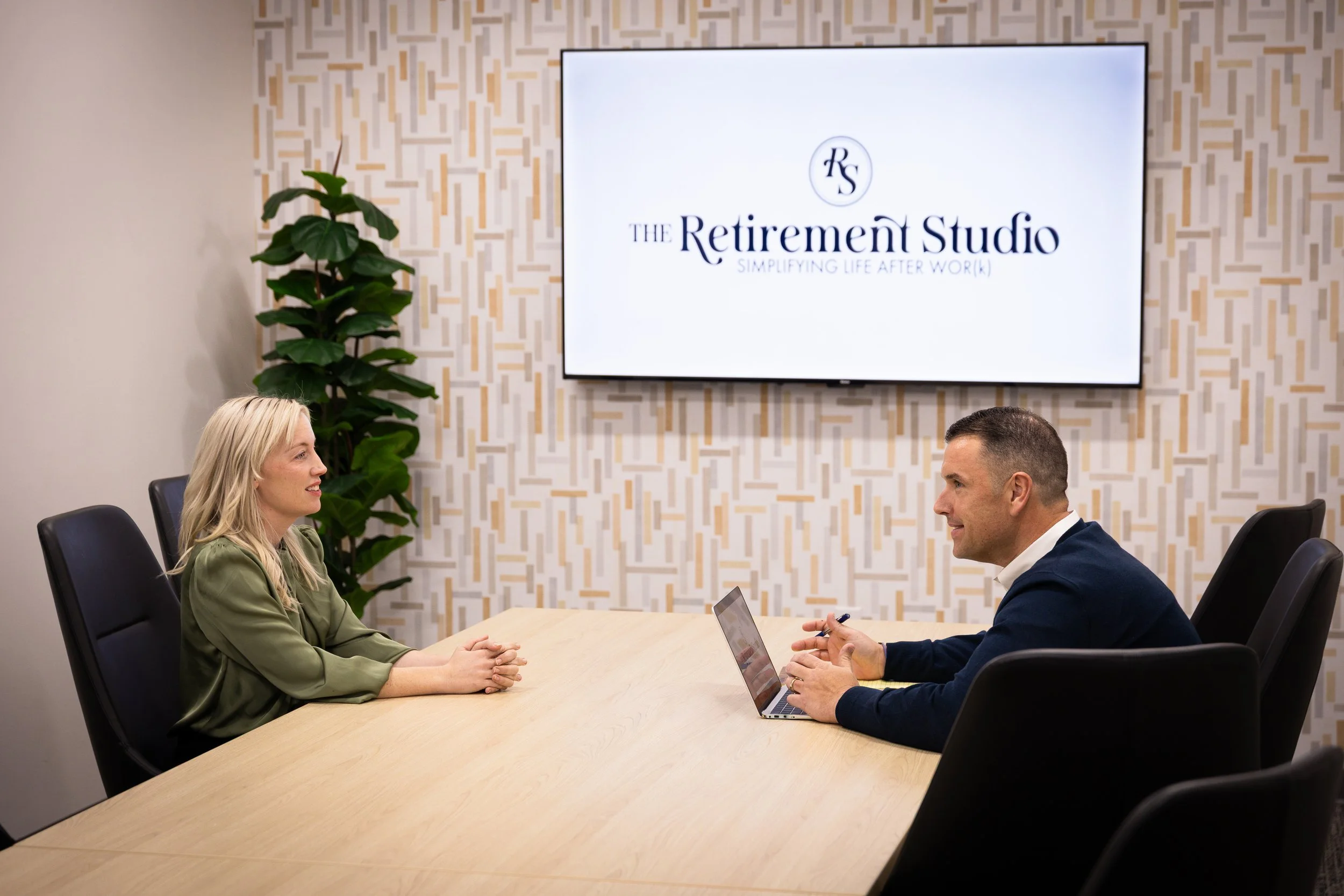 Two people sitting across from each other at a conference table in a meeting room, engaged in conversation. A large screen behind them displays the logo and name "The Retirement Studio" with the tagline "Simplifying Life After Work." A woman with blonde hair wearing a green blazer and a man with short hair wearing a navy blazer are present. The room has a modern decor with a patterned wallpaper and a potted plant in the corner.