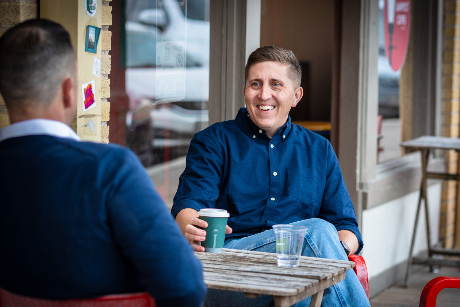 Two men sitting outdoors at a small wooden table, engaging in conversation. One is smiling, holding a coffee cup, while the other, seen from the back, wears a navy sweater. They are at a building with large windows and brick and wooden exterior elements.