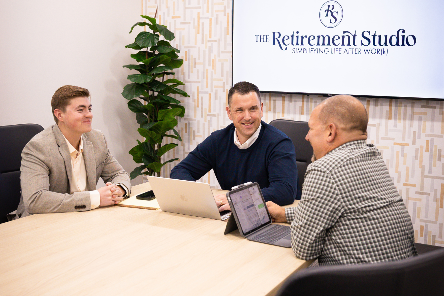 Three men sitting at a conference table during a meeting, smiling and talking. Behind them is a large screen displaying 'The Retirement Studio' logo and tagline.