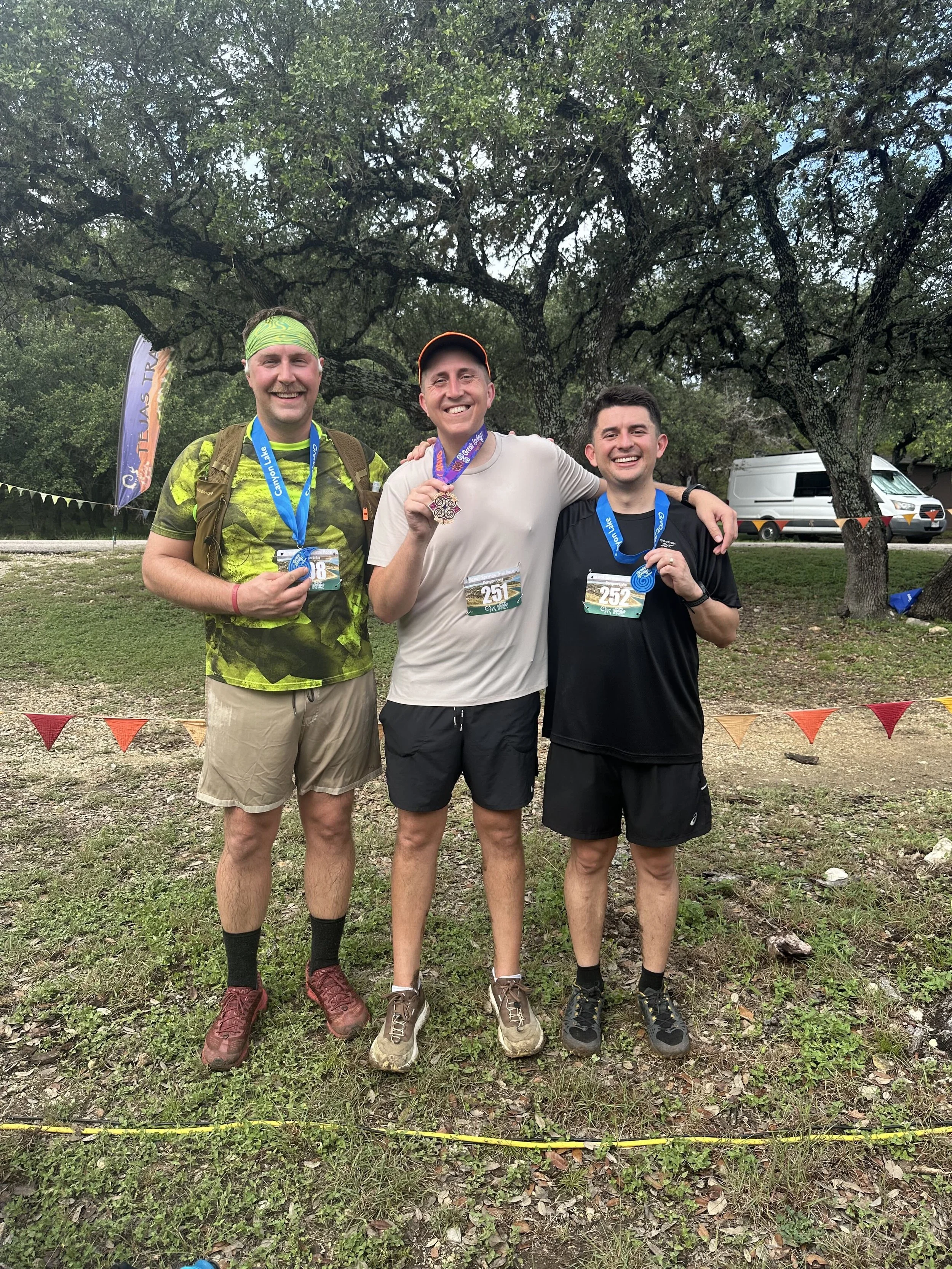Three men celebrating after a race, standing outdoors with trees and a white van in the background, holding medals and wearing race bibs.