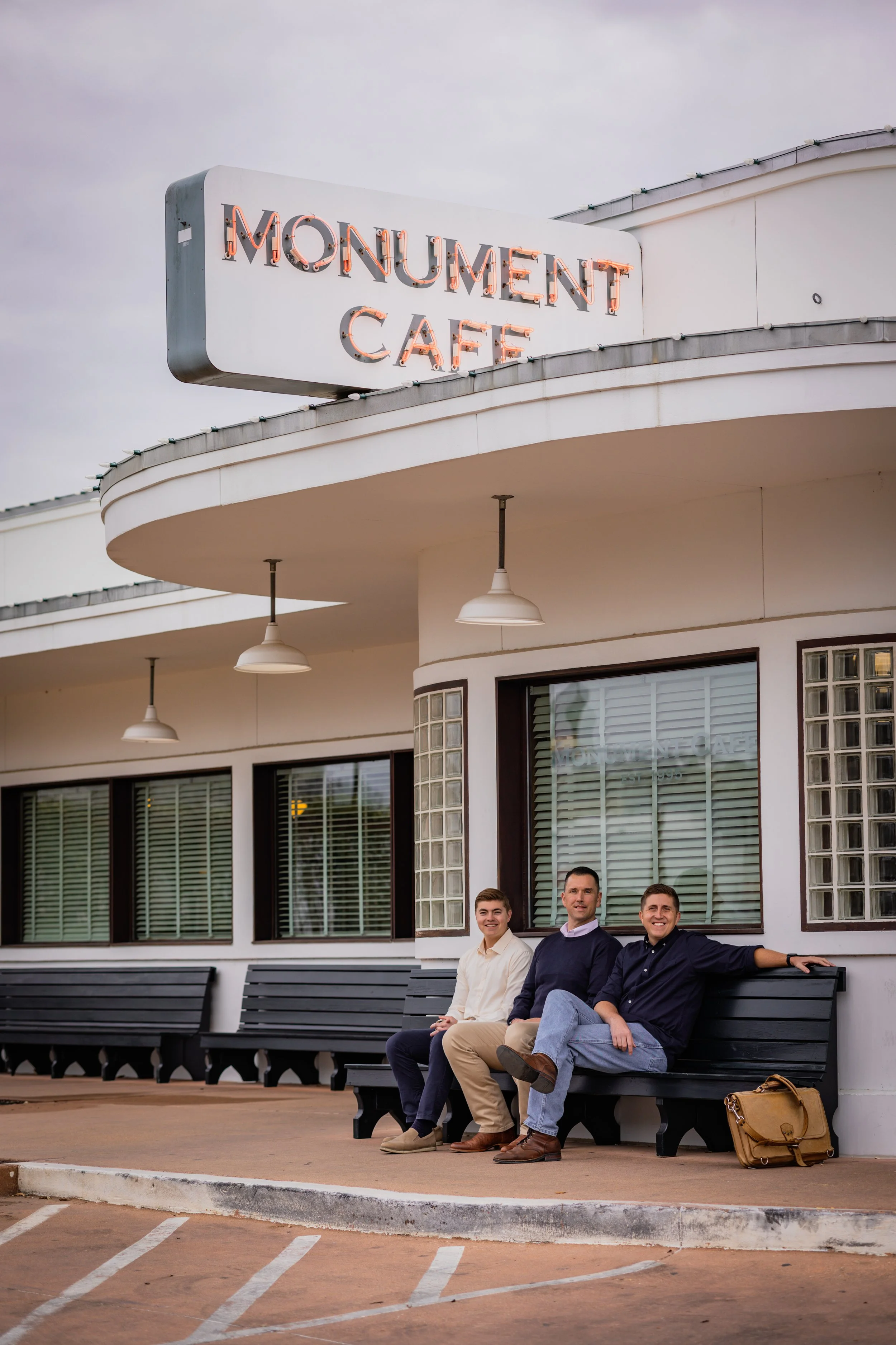 Three young men sitting on a black bench in front of a cafe with a neon sign that reads 'Monument Cafe'. They are smiling and relaxed, with a backpack on the ground beside them.