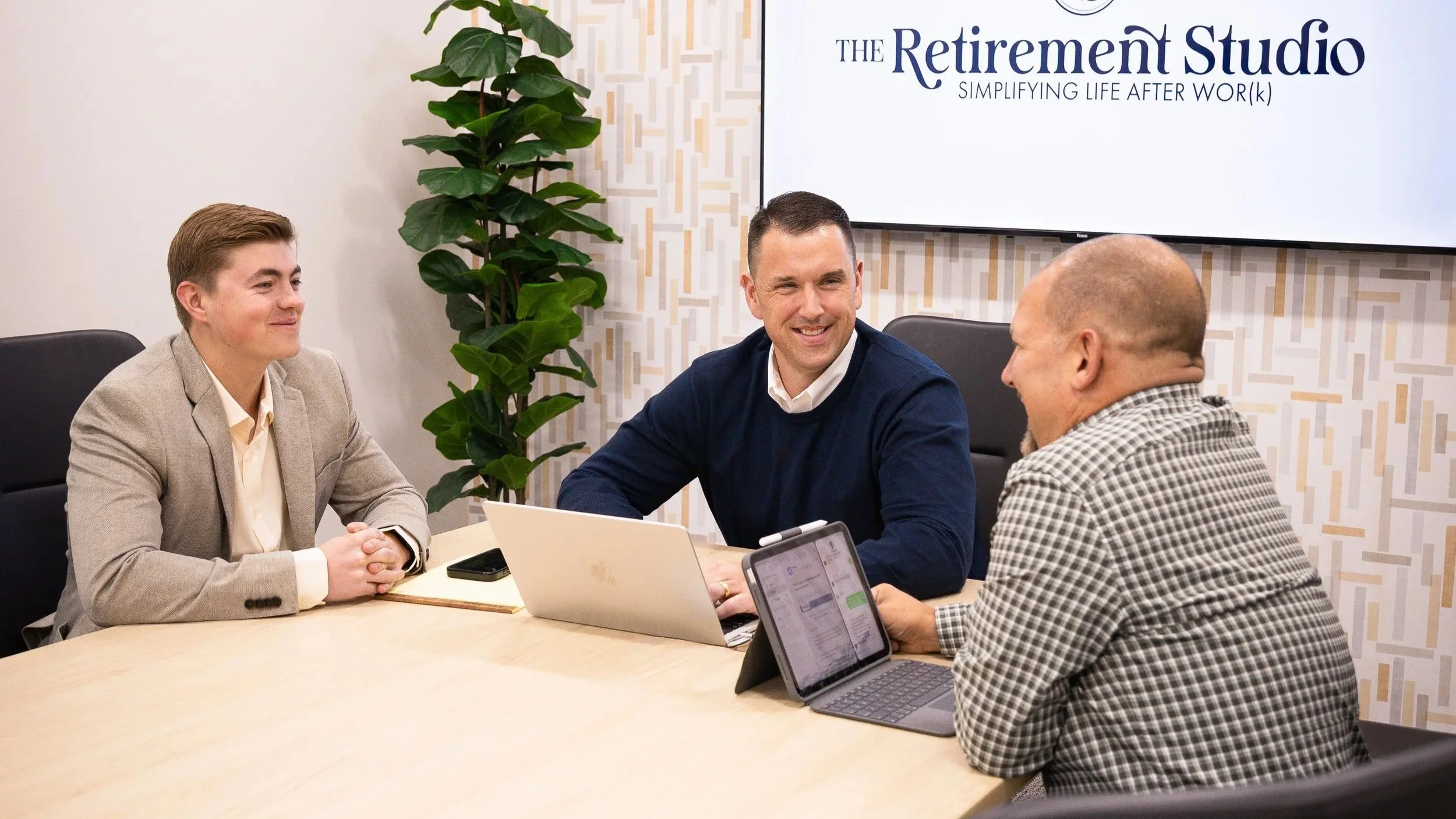 Four men sitting around a conference table, engaged in discussion, with laptops and tablets in front of them. A large screen behind them displays "The Retirement Studio - Simplifying Life After Work." There is a tall potted plant next to the screen and patterned wallpaper on the wall.