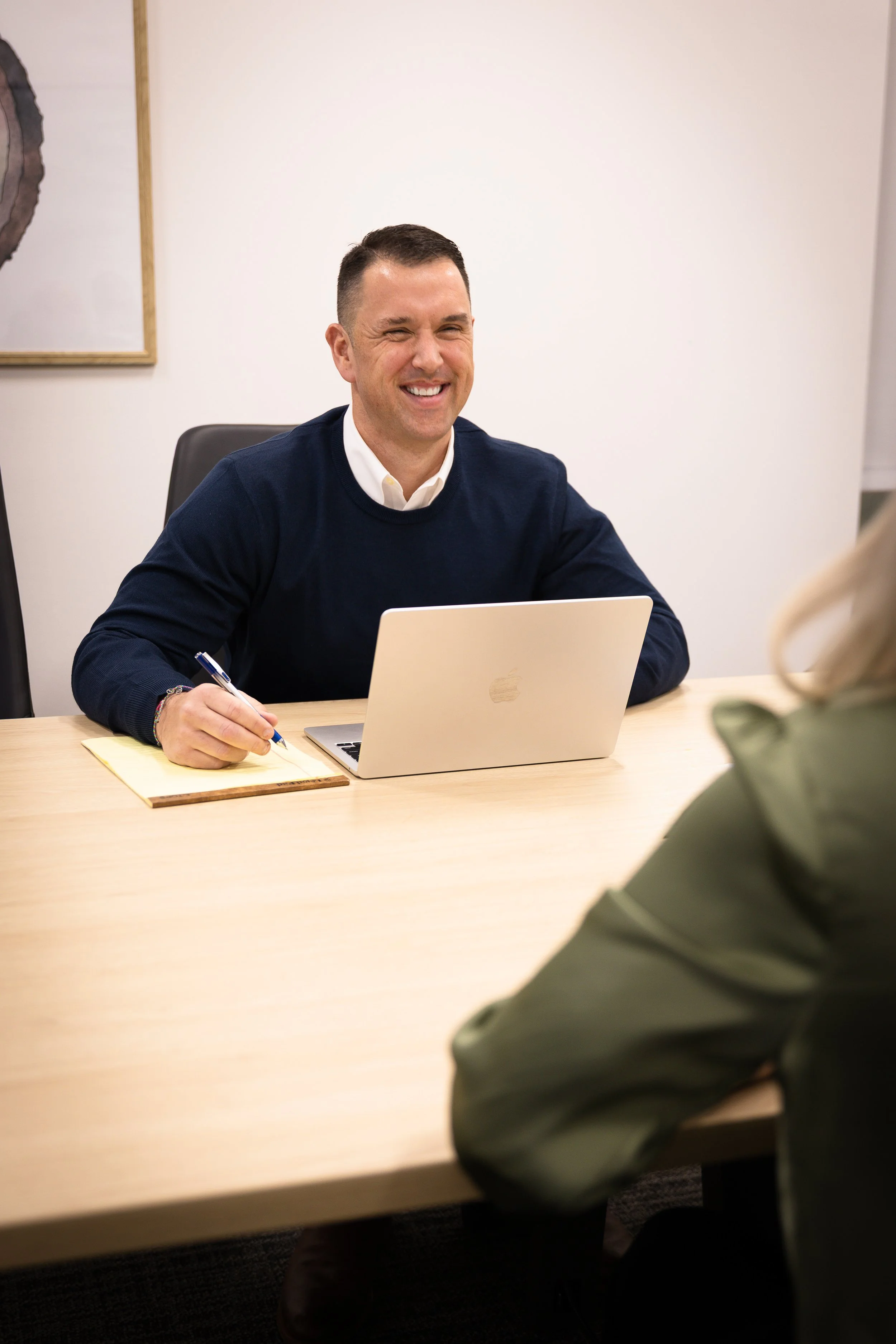 Man with short dark hair, wearing a navy blue sweater, smiling while sitting across a table from a person in a green jacket in a meeting room.
