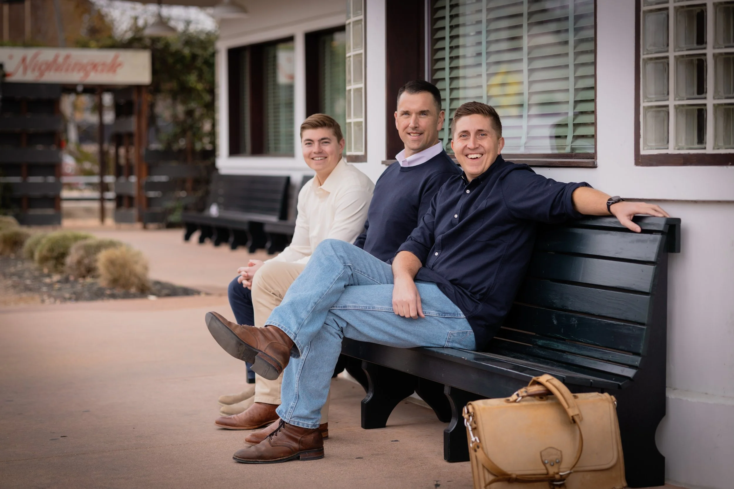 Three men sitting on a bench outside, smiling, with a beige briefcase on the ground near them.