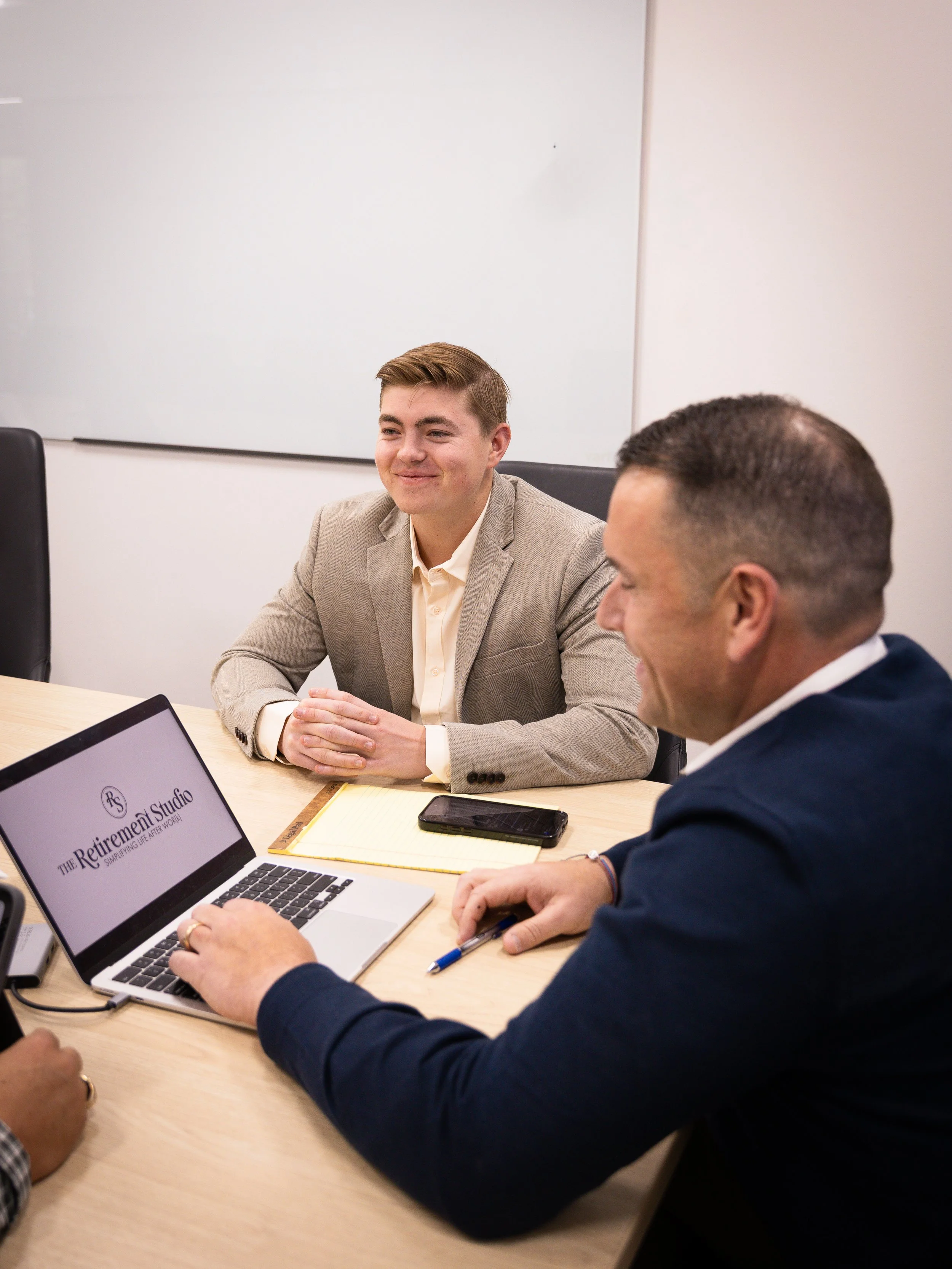 Two men in business attire sitting at a conference table in an office, engaging in a discussion. One man is smiling, the other is taking notes on his laptop, which displays the logo for 'The Retirement Studio'.