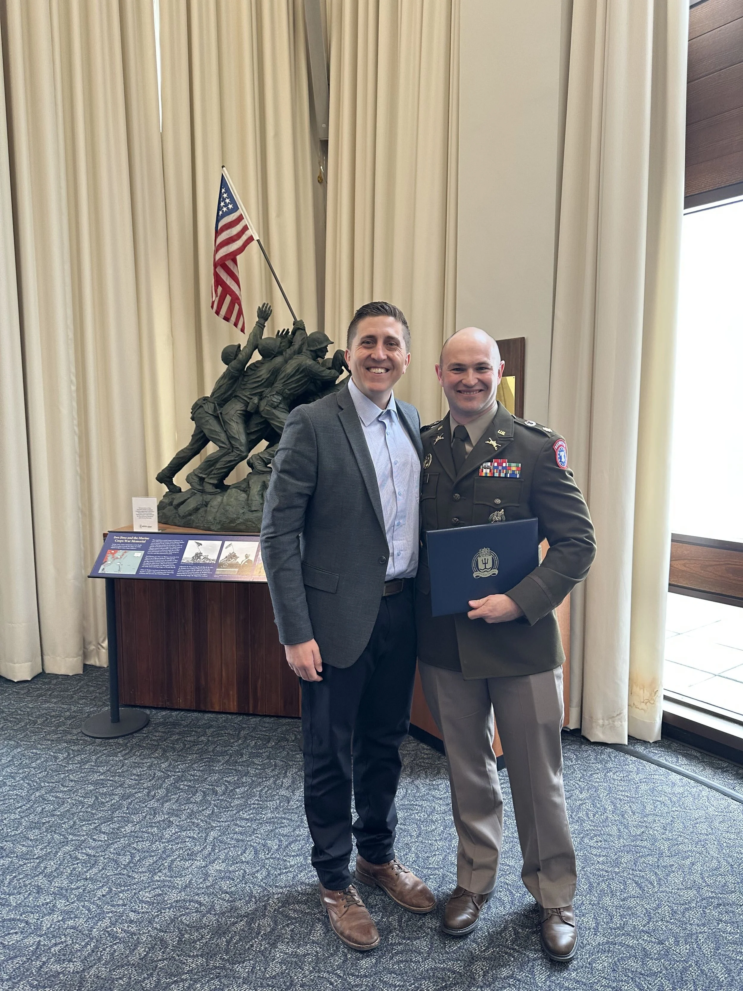 Two men standing together, smiling, in front of a memorial sculpture of soldiers with an American flag. One man is in civilian attire, and the other in military uniform.