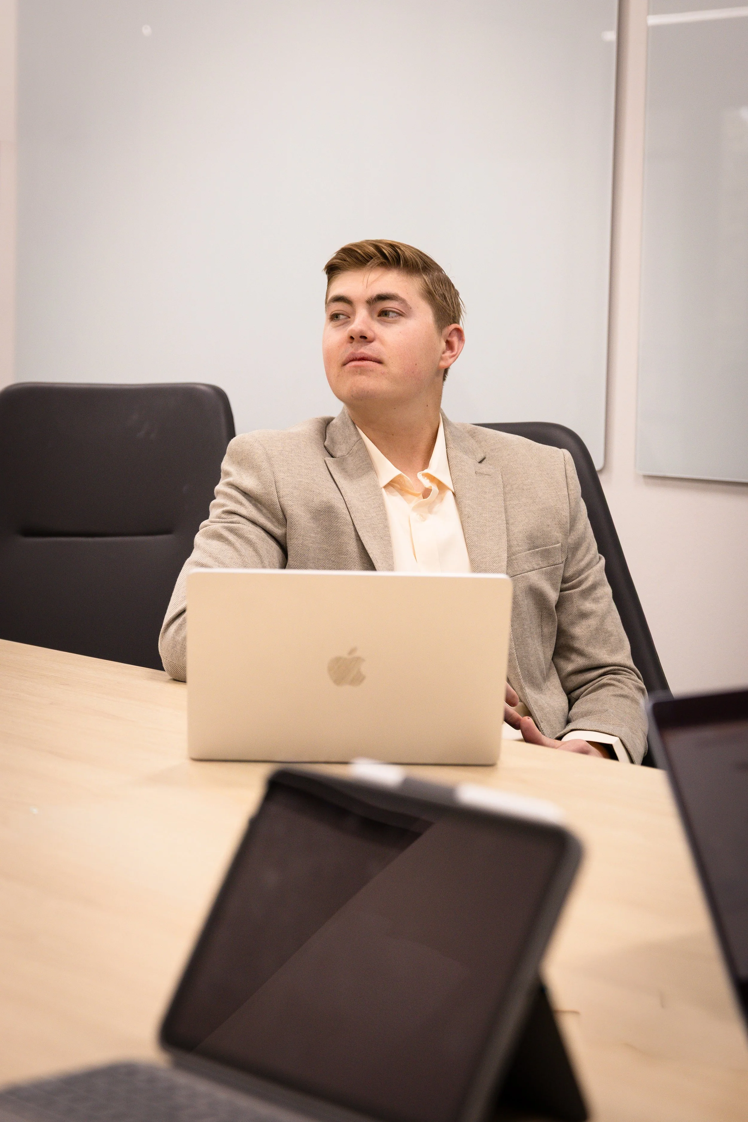 A young man in a beige blazer and white shirt sitting at a conference table with a silver MacBook in front of him, in a modern office setting.