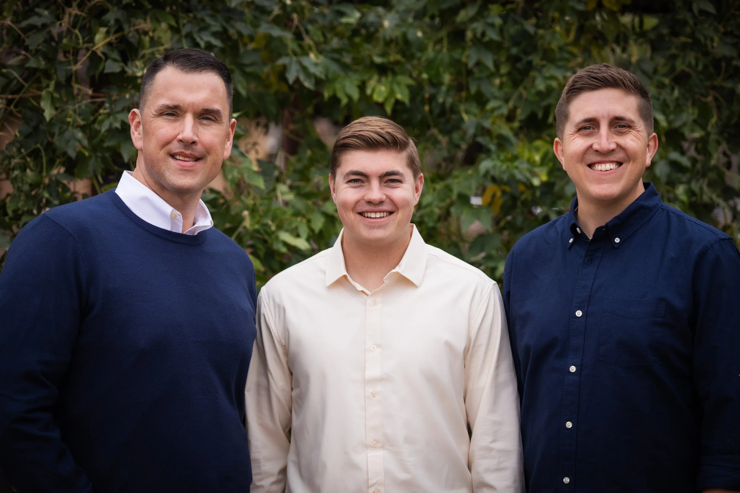 Three men standing outdoors in front of greenery, smiling at the camera, dressed in collared shirts and sweaters.
