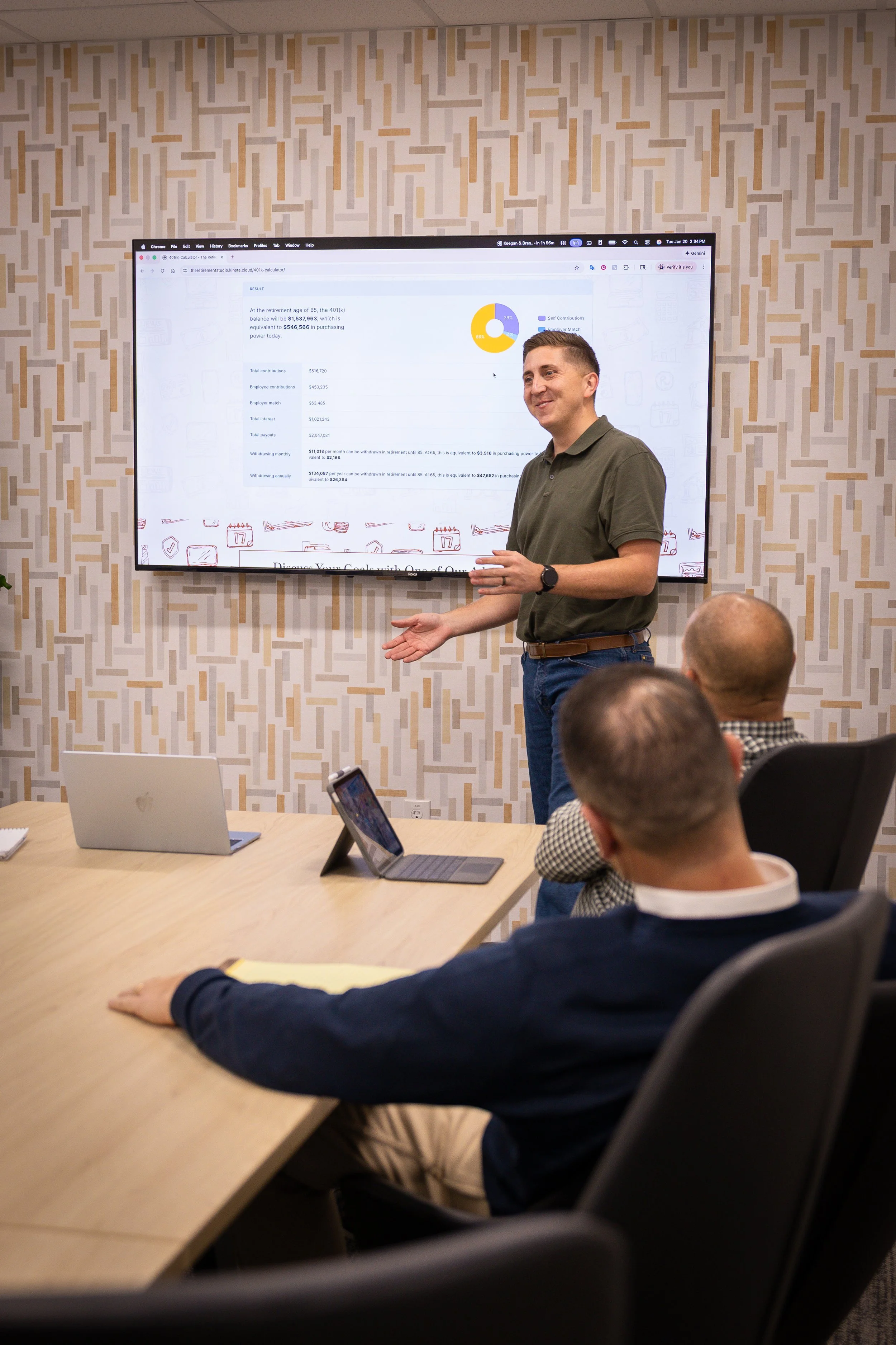 A man in a green polo shirt giving a presentation in a conference room with patterned wallpaper. He is standing in front of a large screen displaying a financial chart and text, with an audience seated at a wooden table using laptops and tablets.