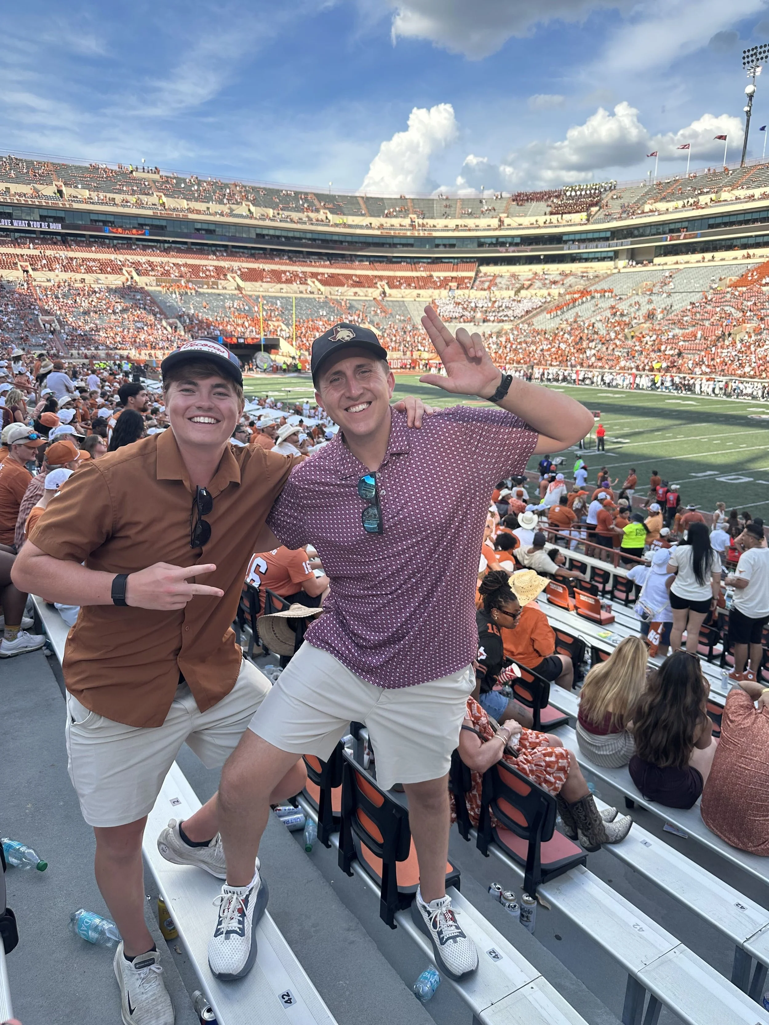 Two young men at a football stadium, smiling and posing for the photo with the stands and crowd in the background. They are wearing casual clothing and hats, and are standing on stadium seats.