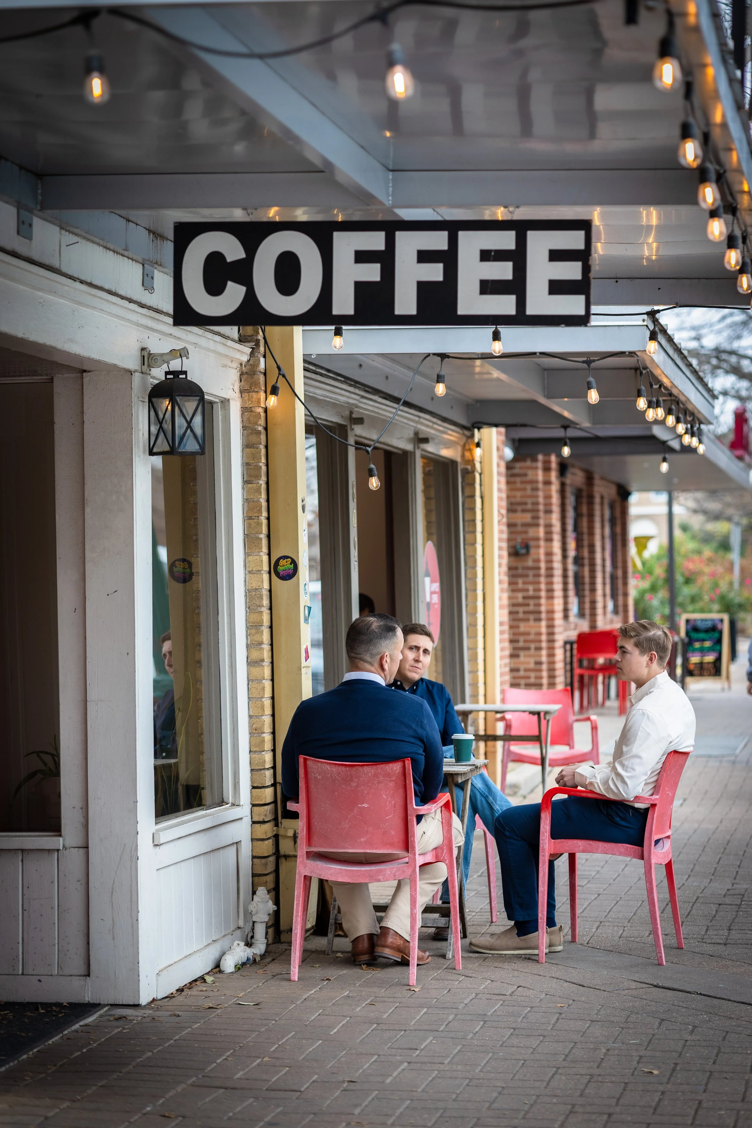Three men sitting outside a cafe under a sign that says "Coffee," with pink chairs and string lights overhead. One man is wearing a white shirt, and two are wearing dark blue suits.