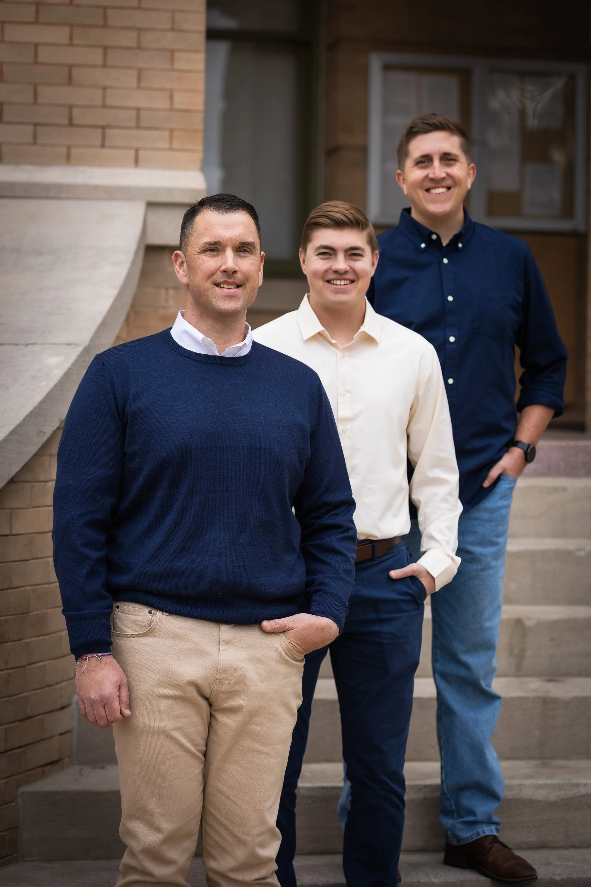 Three smiling men standing in front of a brick and stone building, dressed in casual business attire.
