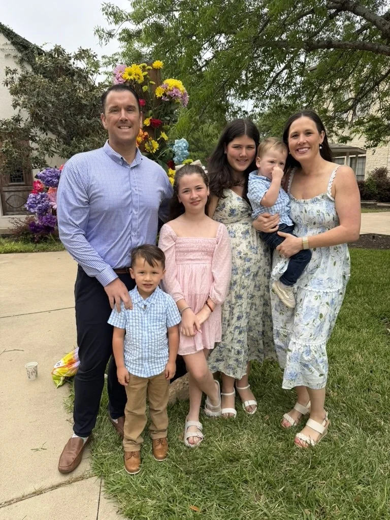A family of six posing outdoors in front of a flower arrangement, standing on a lawn and sidewalk with trees and buildings in the background.