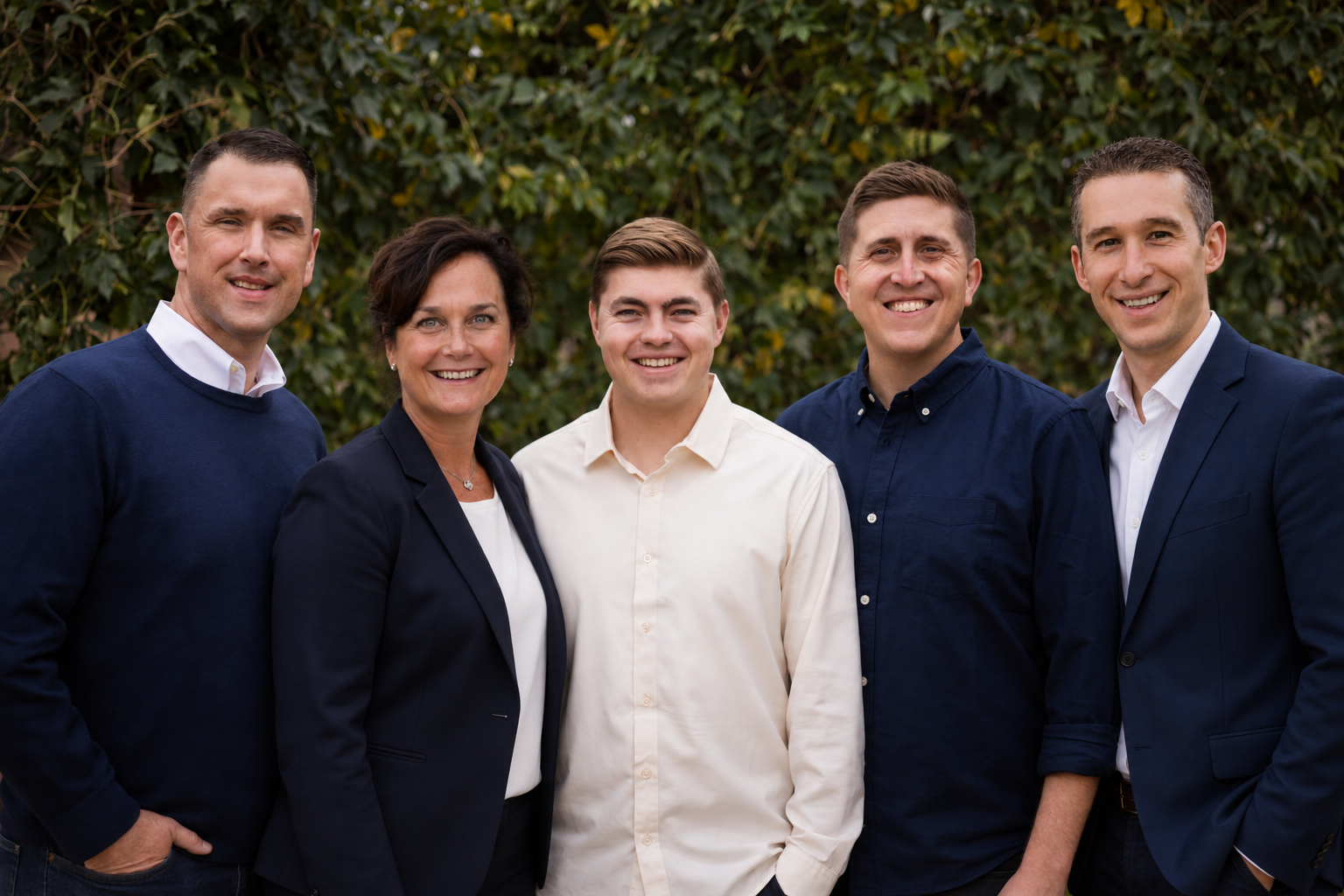 Group of five professionally dressed people standing outdoors in front of greenery, smiling at the camera.