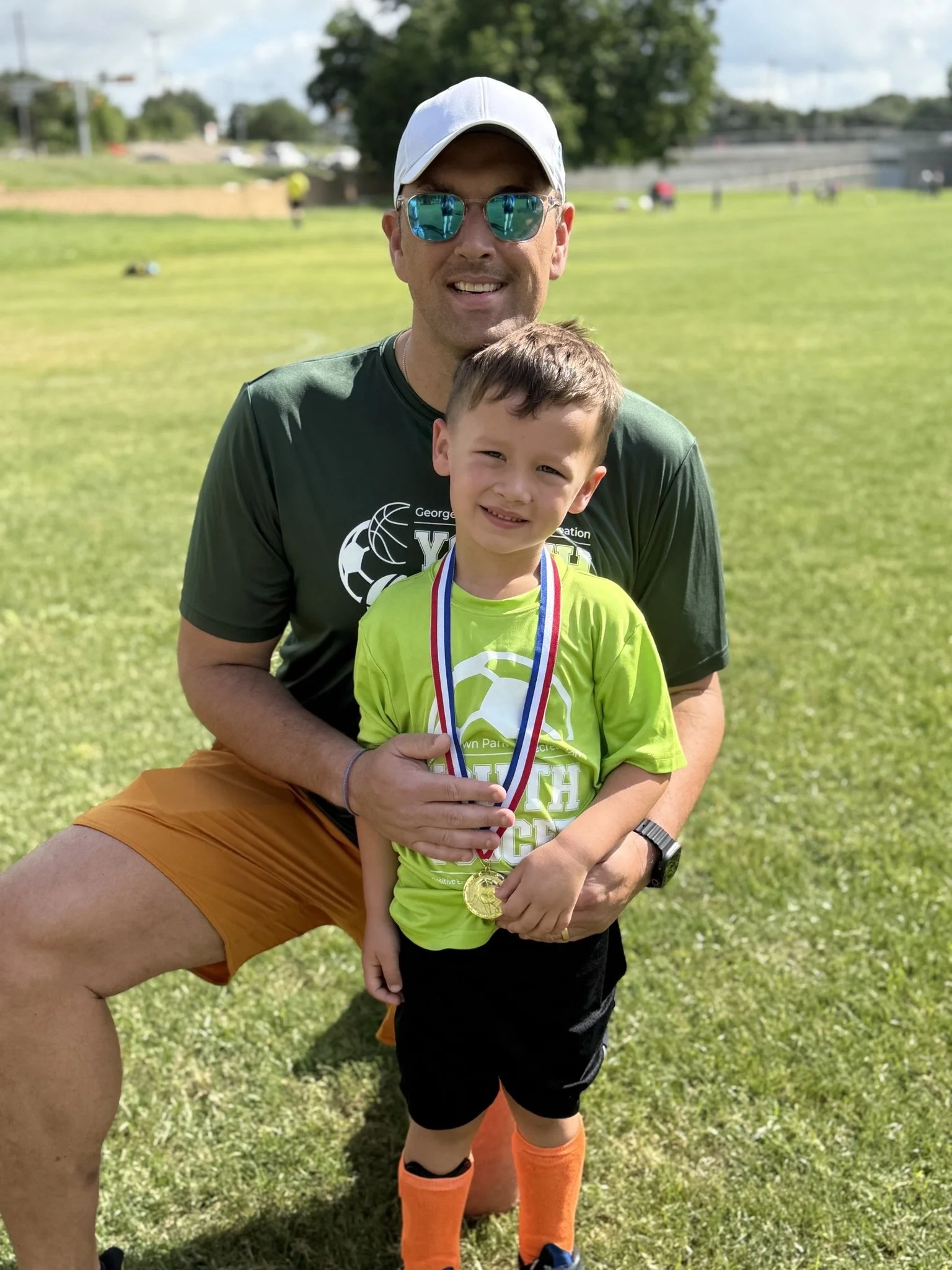 A man and a young boy standing on a grassy field, celebrating a sports event. The boy is wearing a neon green shirt with a medal around his neck, and the man is in a black shirt, orange shorts, and sunglasses. The man is kneeling behind the boy, holding him, and both are smiling at the camera.