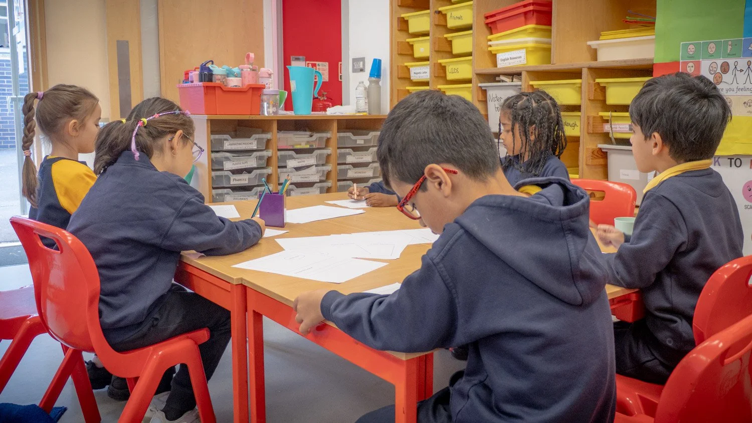 Children sitting at a classroom table, working on papers with pencils, in a colorful classroom with storage bins and educational posters.