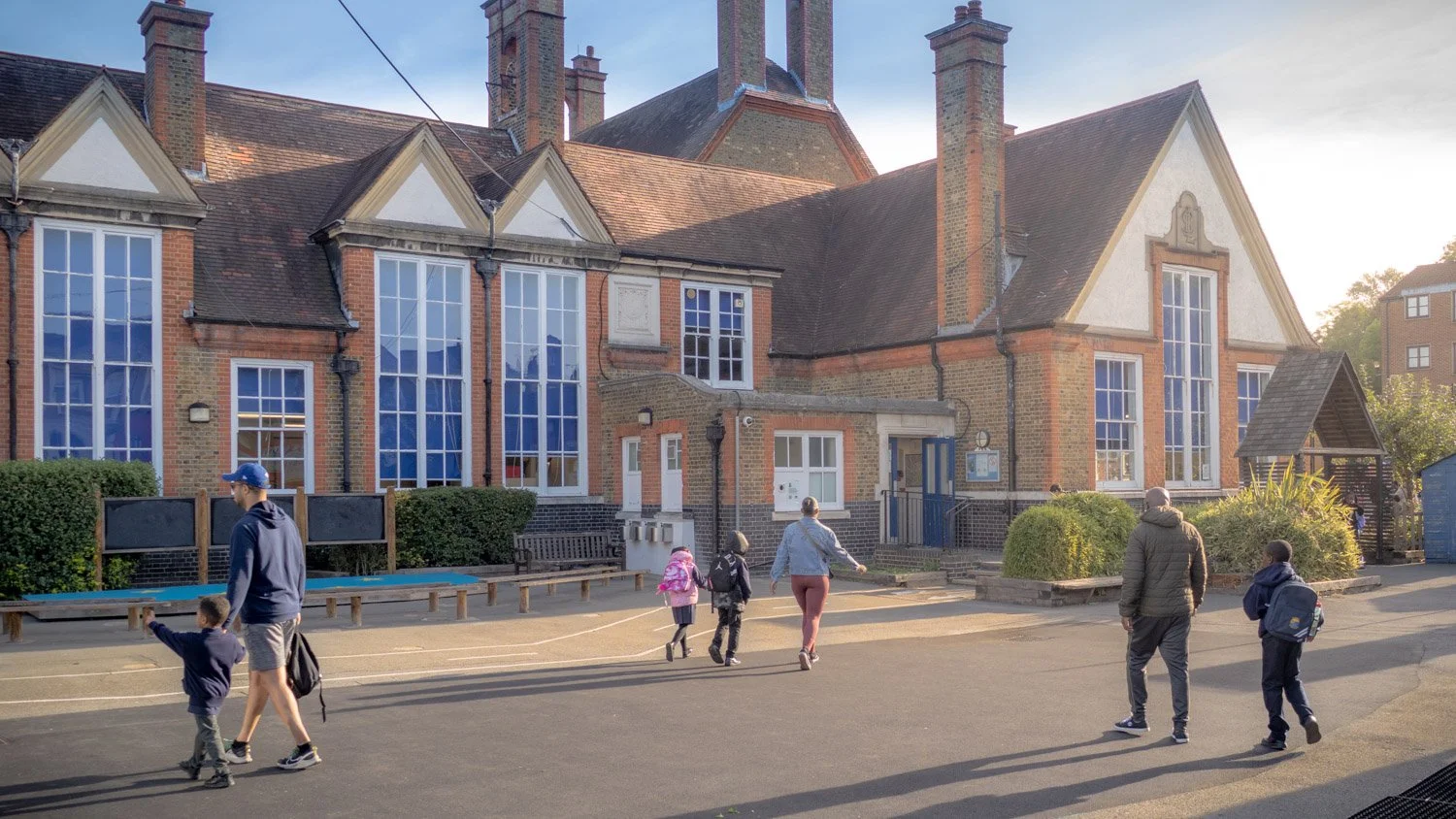 Children and adults walk outside a brick school building with large windows, bushes, and a small wooden structure in the background.