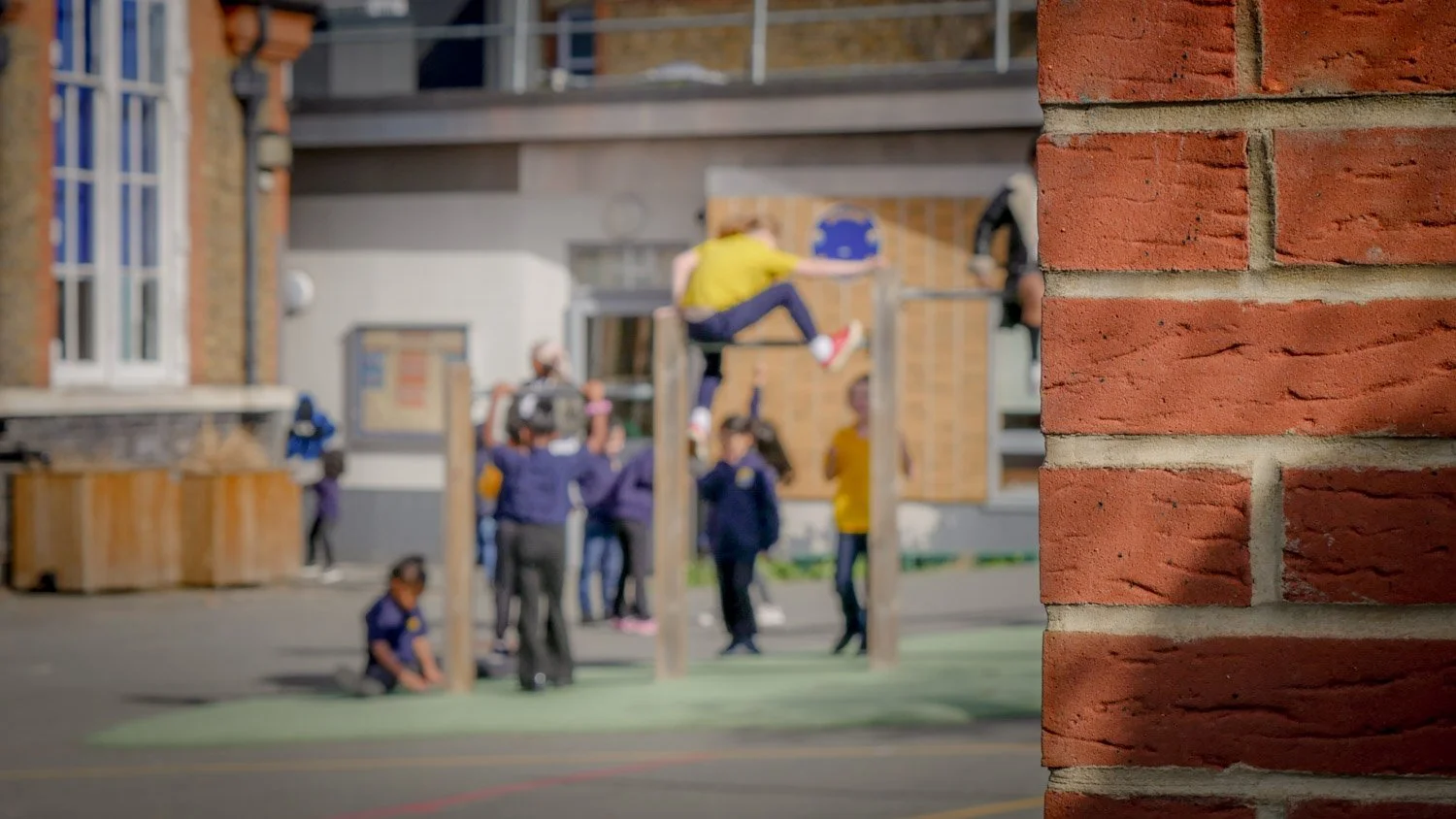 School children playing on outdoor equipment at a playground, with a brick wall in the foreground.