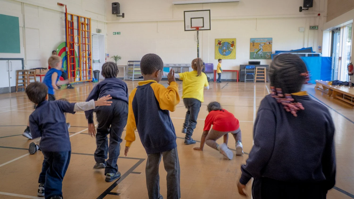 Children in a school gymnasium playing and running with some reaching towards the floor, under a basketball hoop, with bright decor and artwork on the walls.