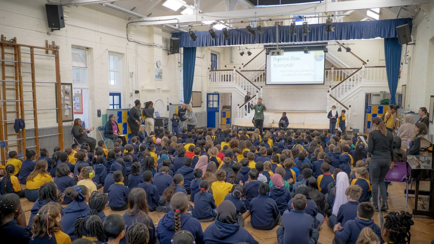 A large group of students seated on the floor in a school auditorium, listening to a speaker on stage. The stage has a large screen displaying a presentation, and there are teachers and staff standing around.