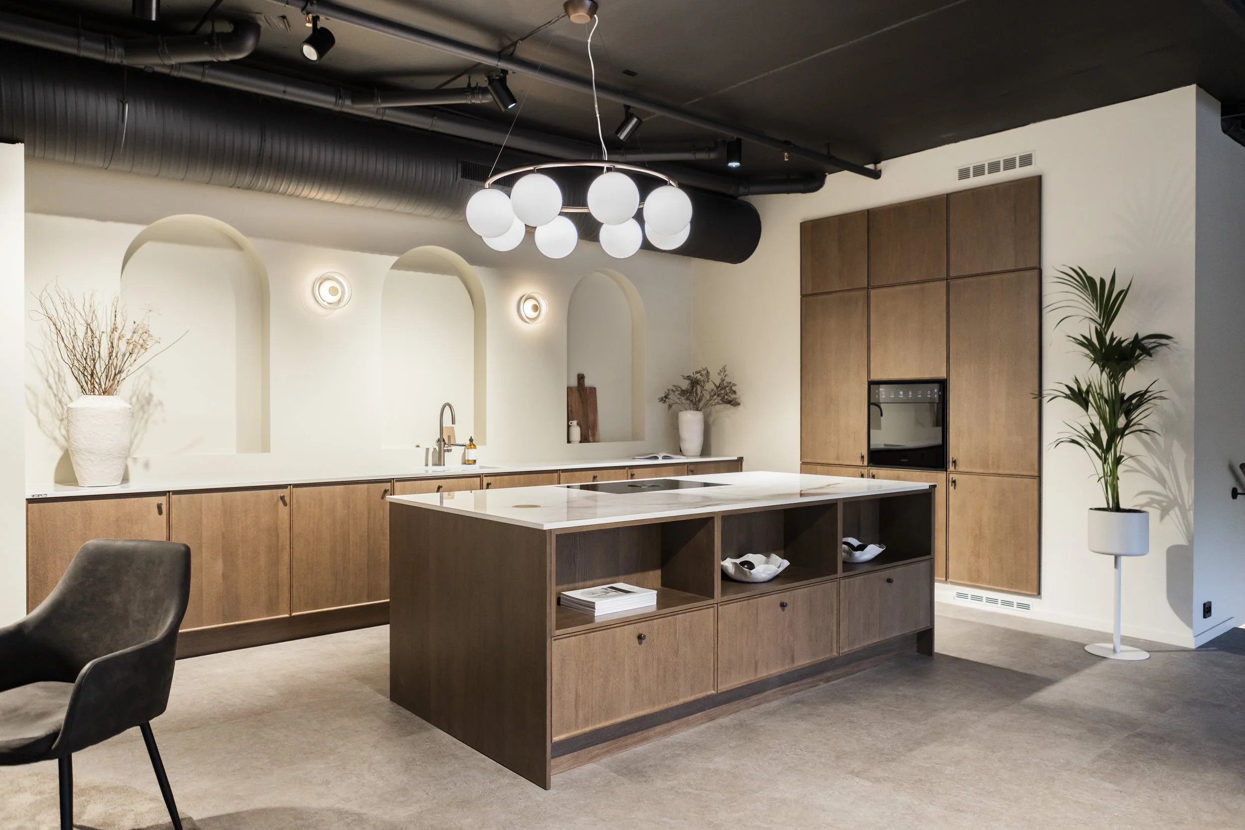 Modern kitchen with wooden cabinets, white island, black chairs, circular pendant light fixture, potted plants, and decorative vases.
